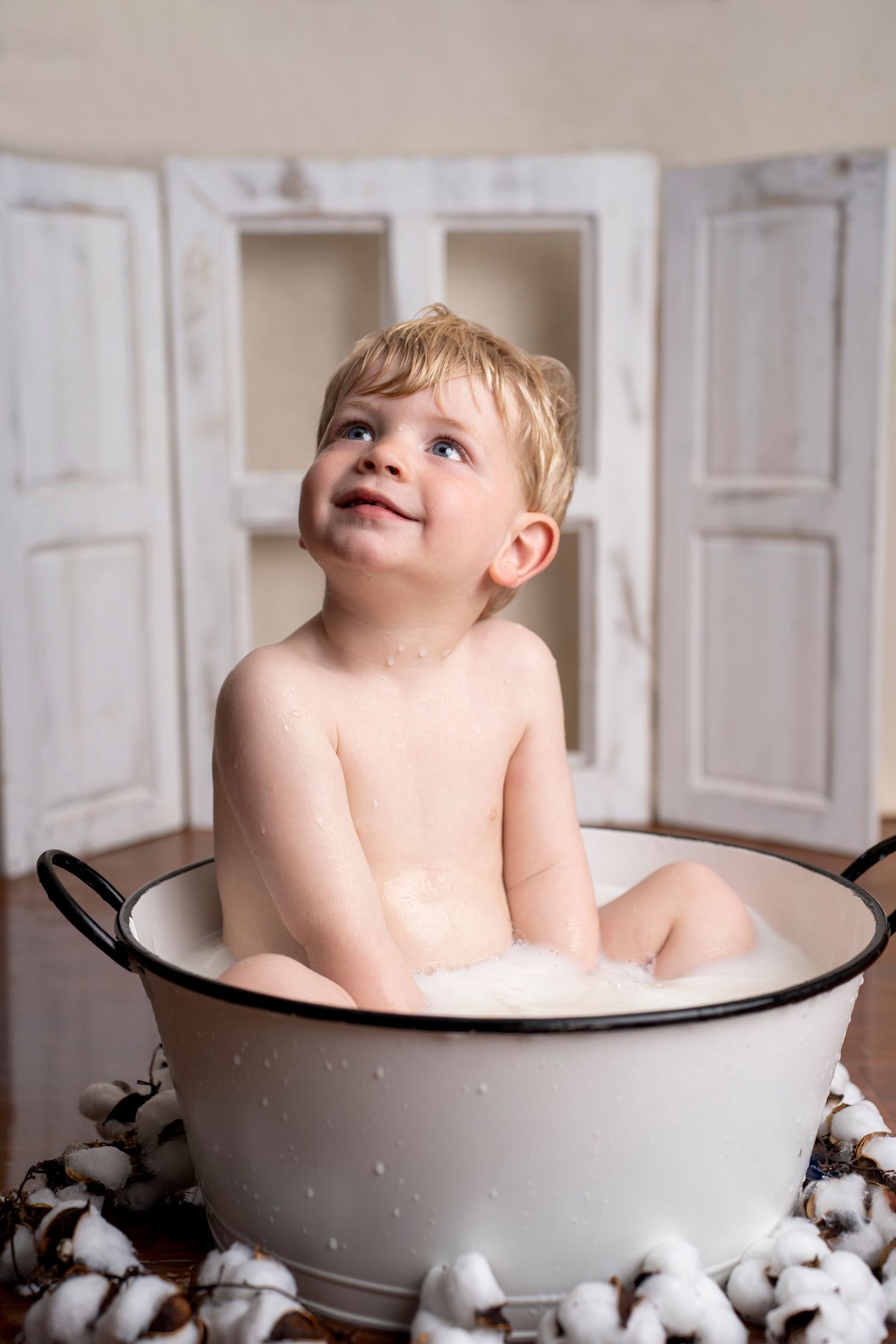 Smiling toddler boy sitting in a white enamel tub with bubble bath, surrounded by cotton bolls.