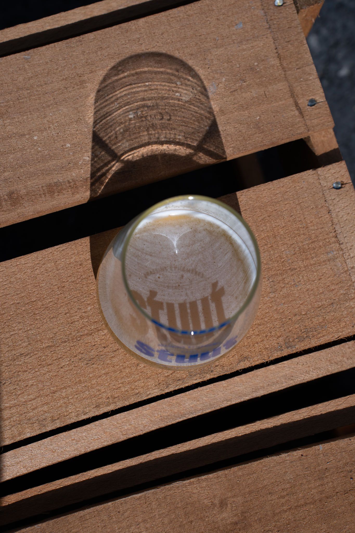 Beer glass casting shadow on wooden deck planks in sunlight.