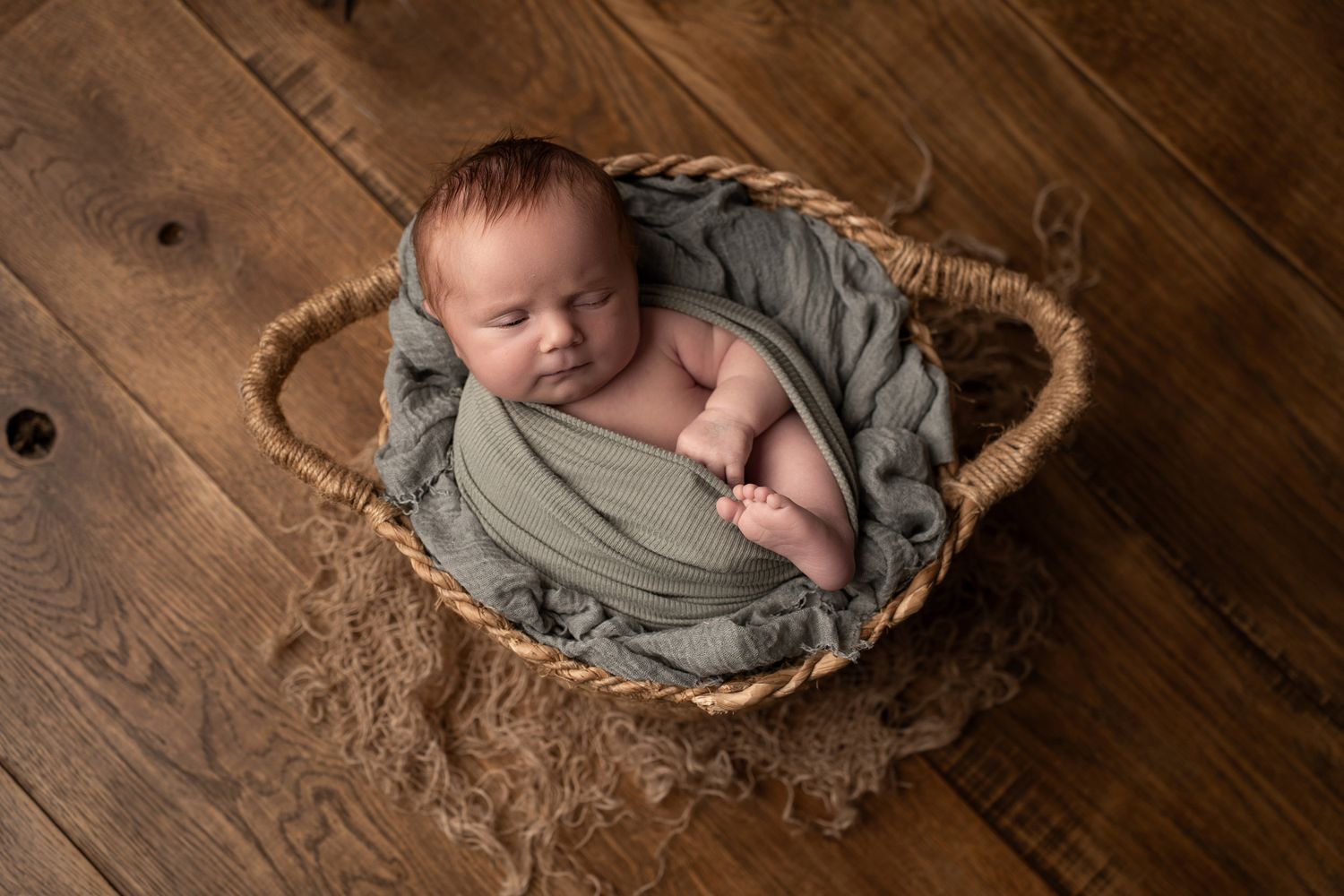 Sleeping newborn wrapped in sage green fabric nestled in rustic basket.