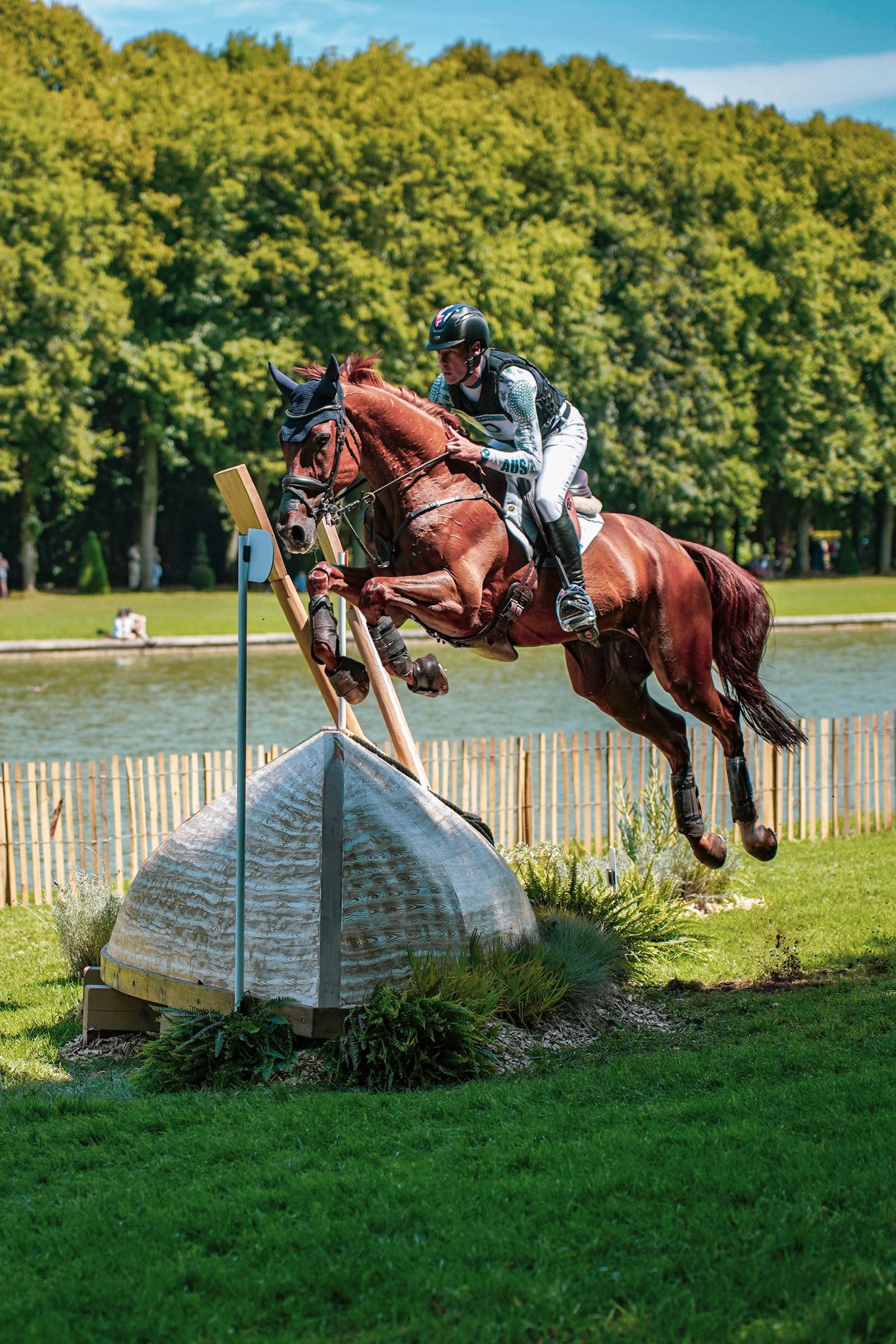 Chestnut horse and rider clearing a large wooden fence jump over water during competition.