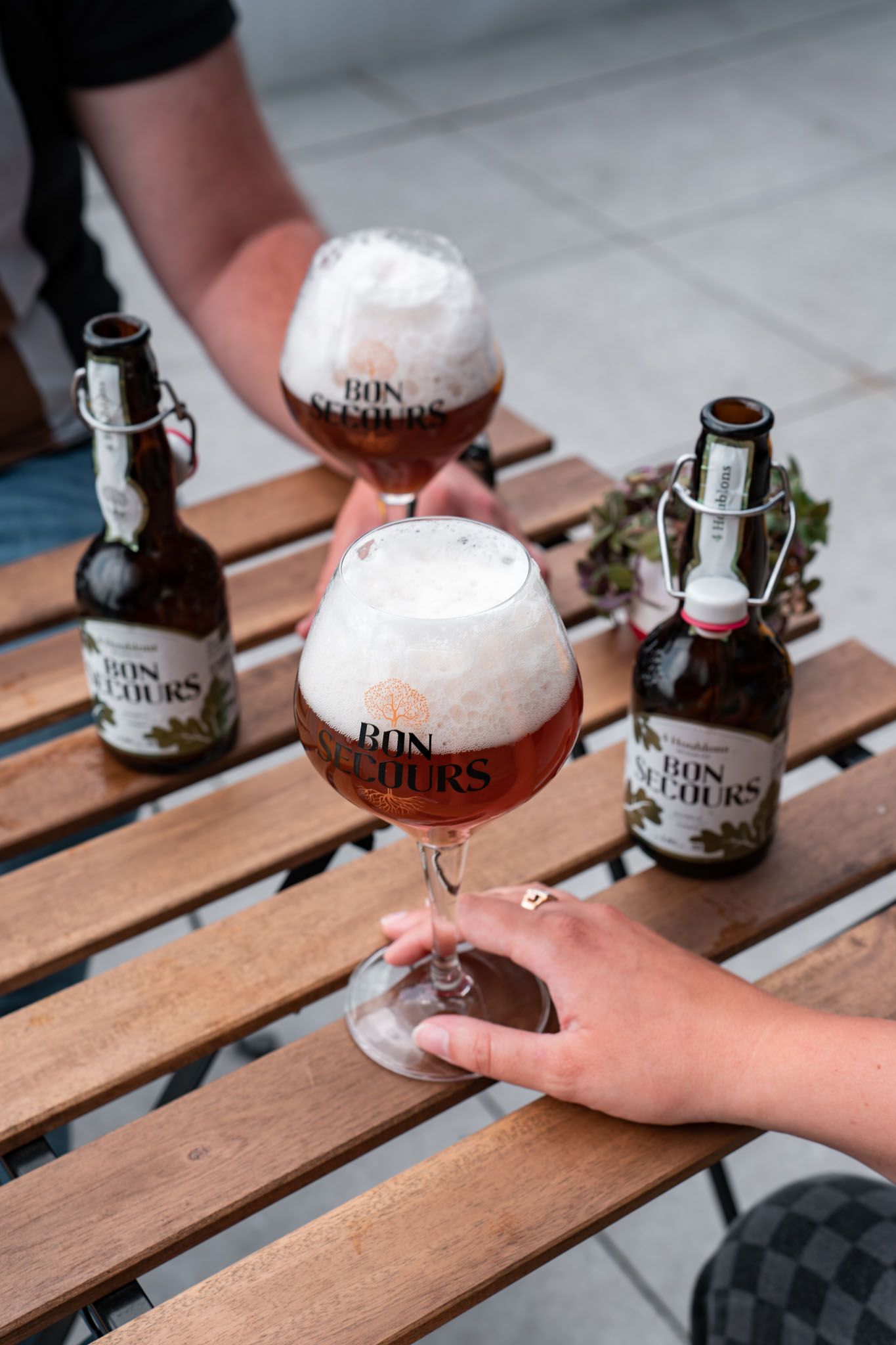 Two glasses of craft beer with frothy heads and bottles on wooden slats.