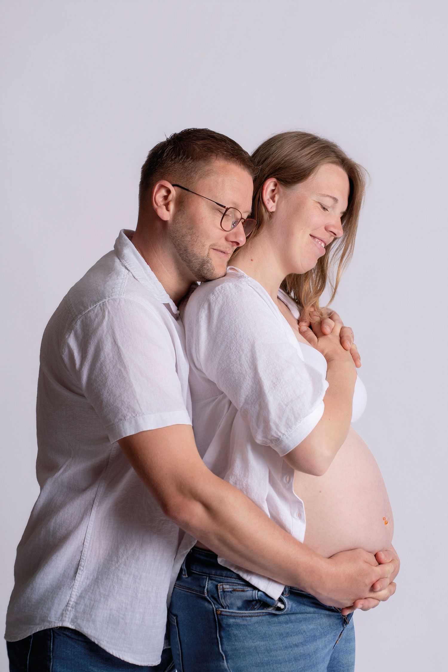 A man tenderly embraces a smiling pregnant woman, both wearing white shirts and jeans against a white background.