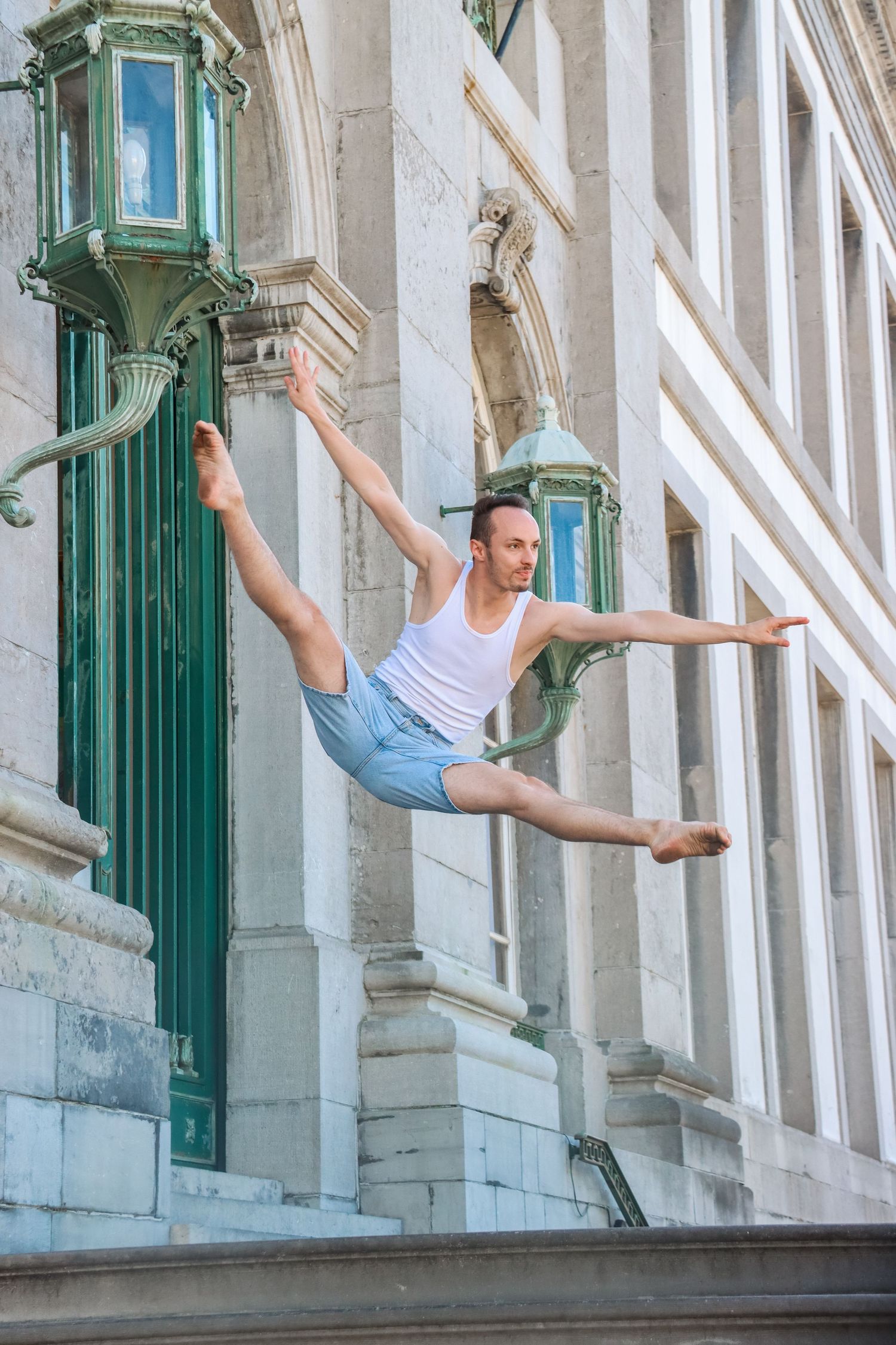 A dancer performs a graceful leap in front of a classical white building with green columns.