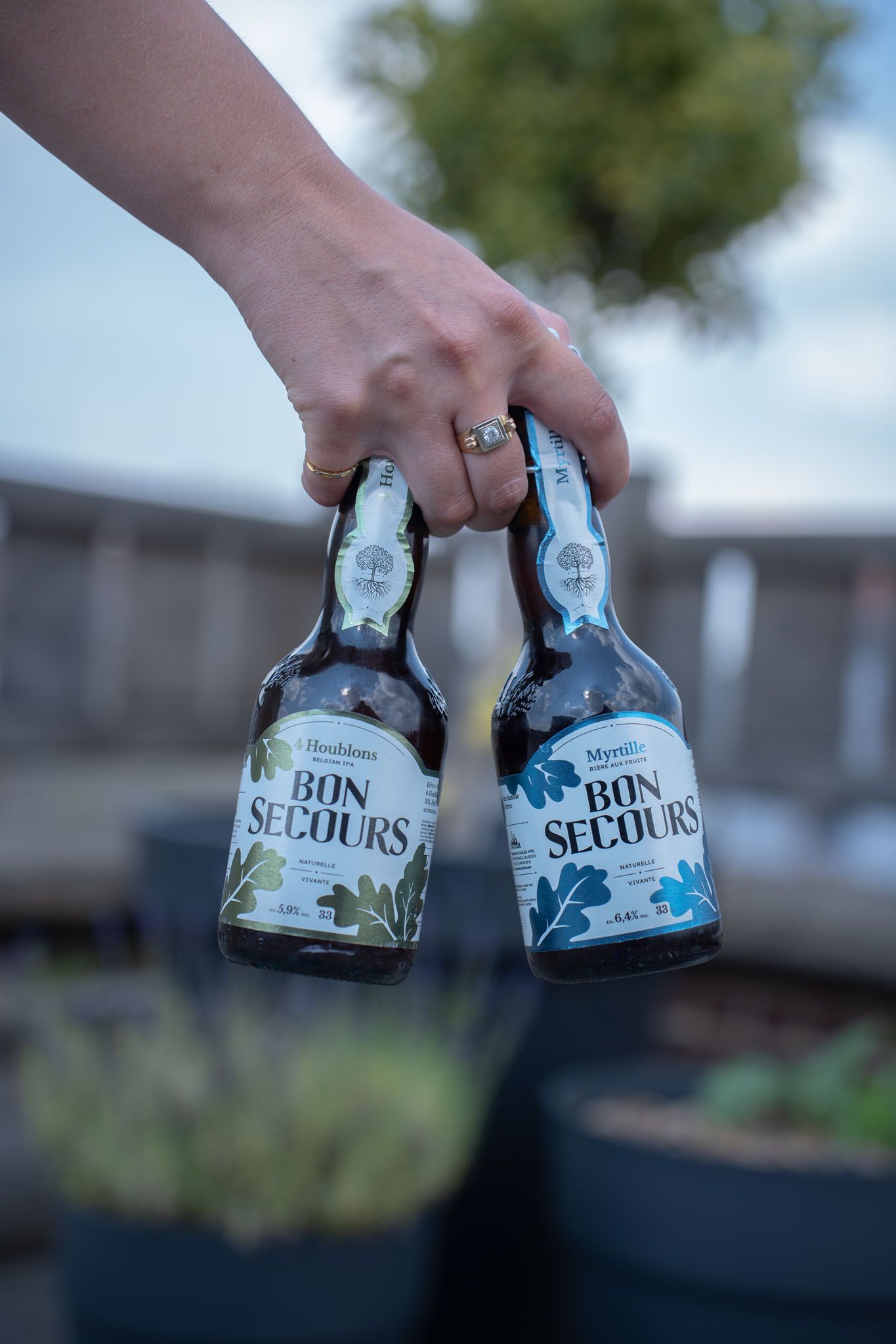 Hand holding two Bon Secours beer bottles against blurred outdoor background.