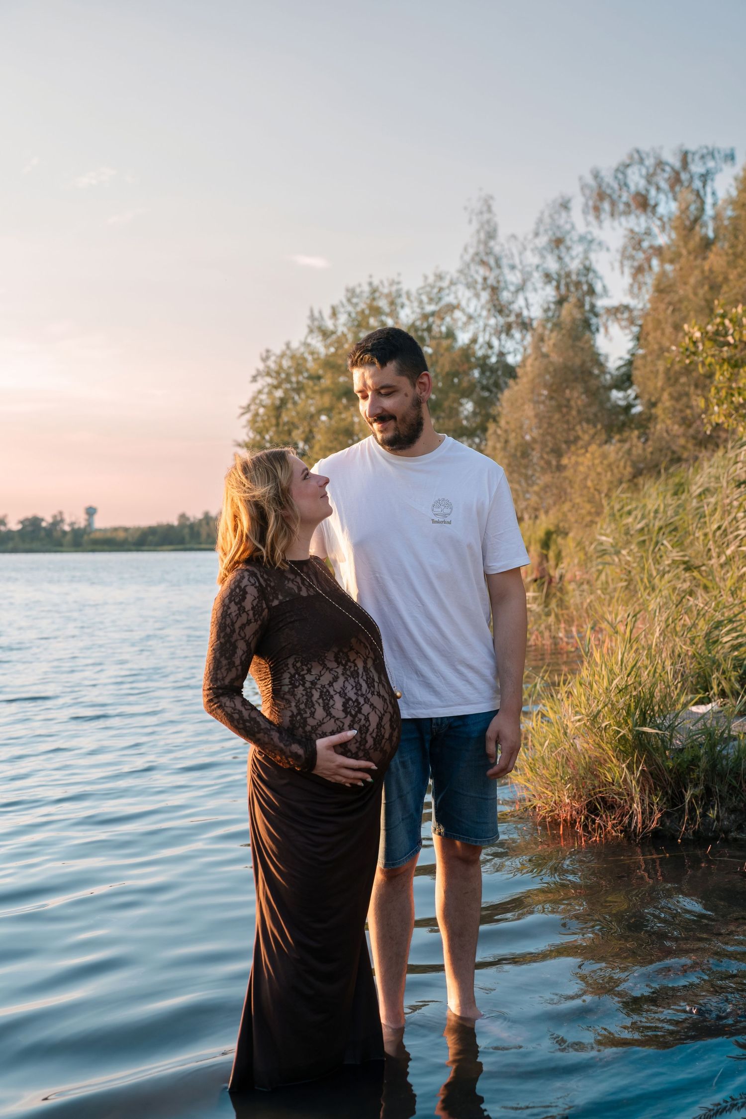 Pregnant couple standing in shallow water at sunset, woman in black lace gown cradling bump, man in white tee and denim shorts.