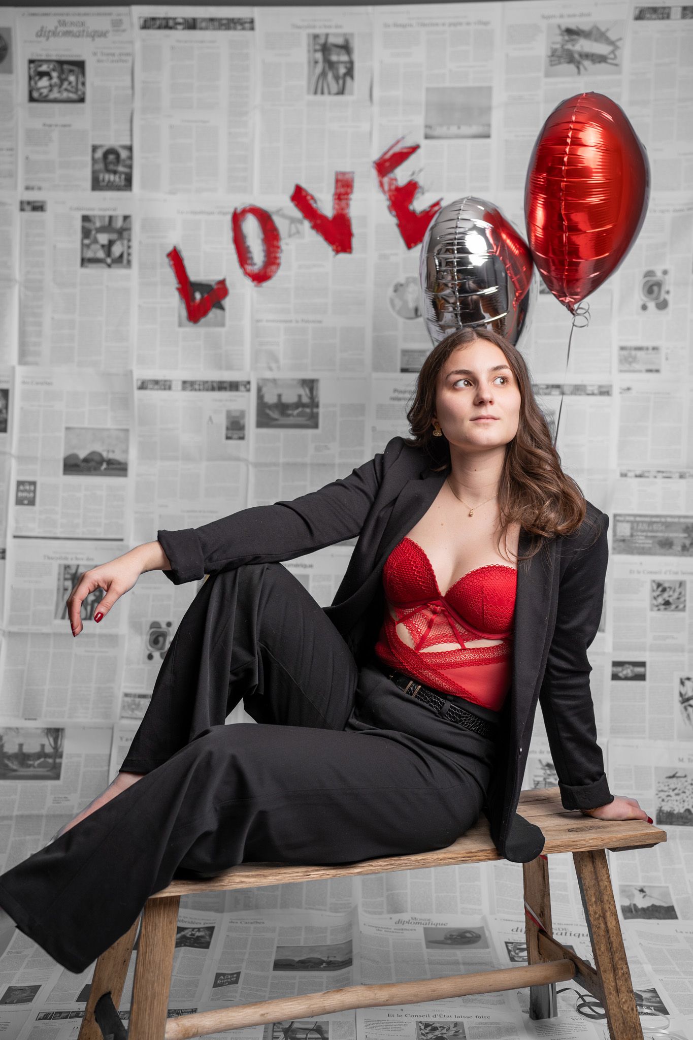 Woman in black suit poses with red heart balloons against newspaper-covered wall in stylish photoshoot.