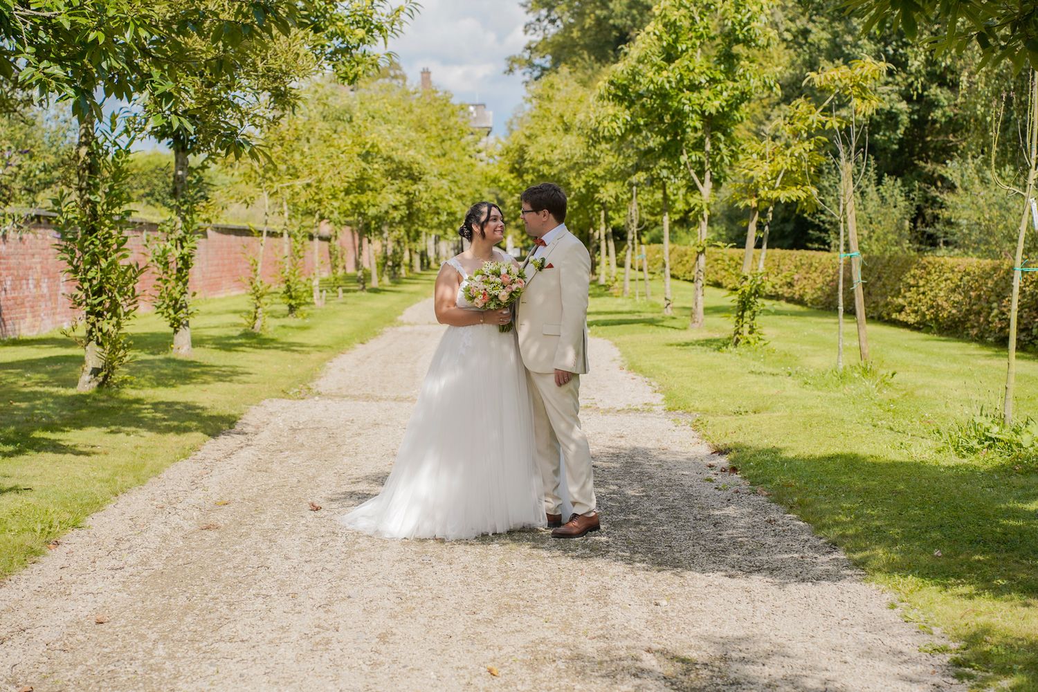 Newlyweds walk together along a tree-lined path in a lush garden setting.