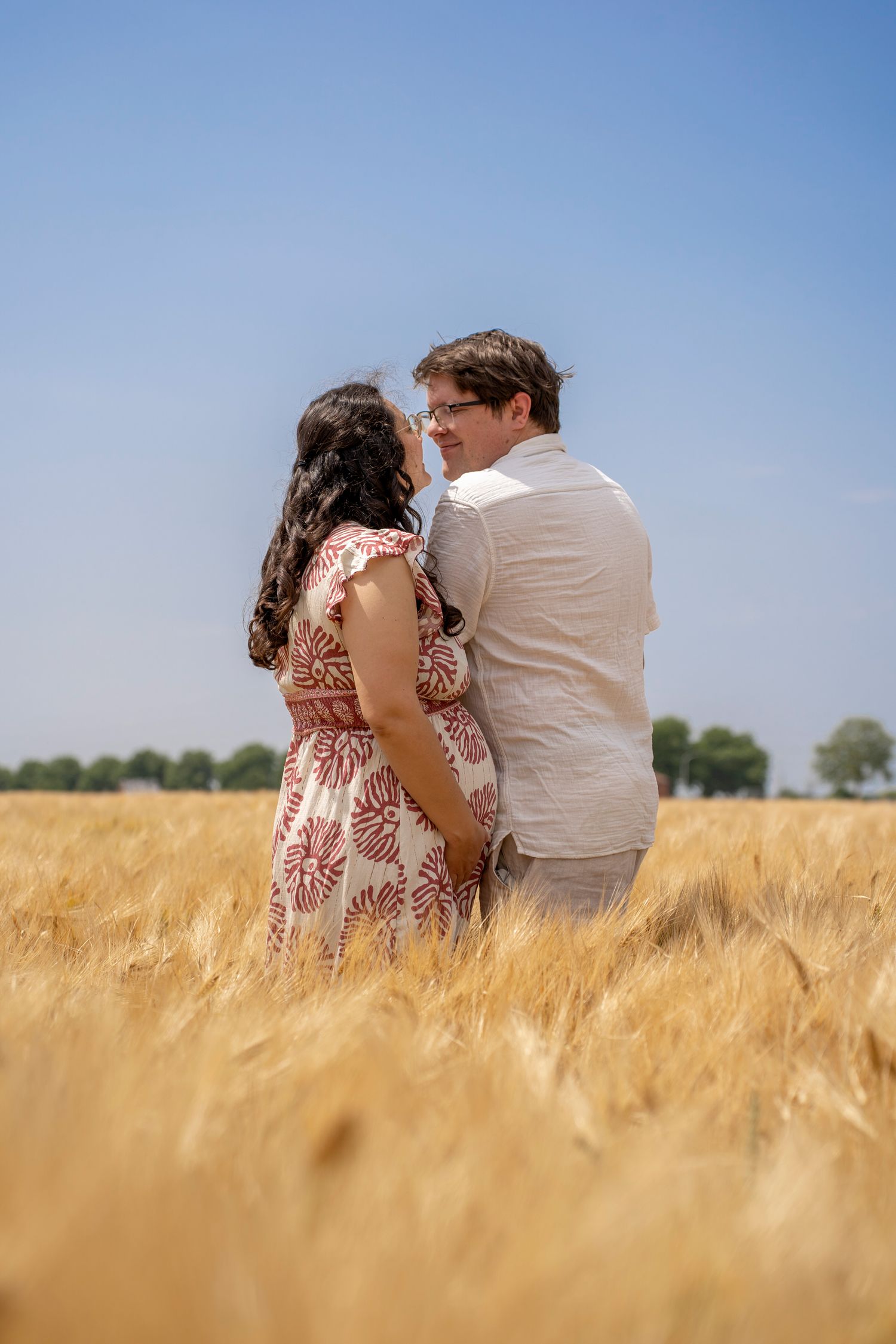 Romantic couple sits close together in golden wheat field under bright blue sky.