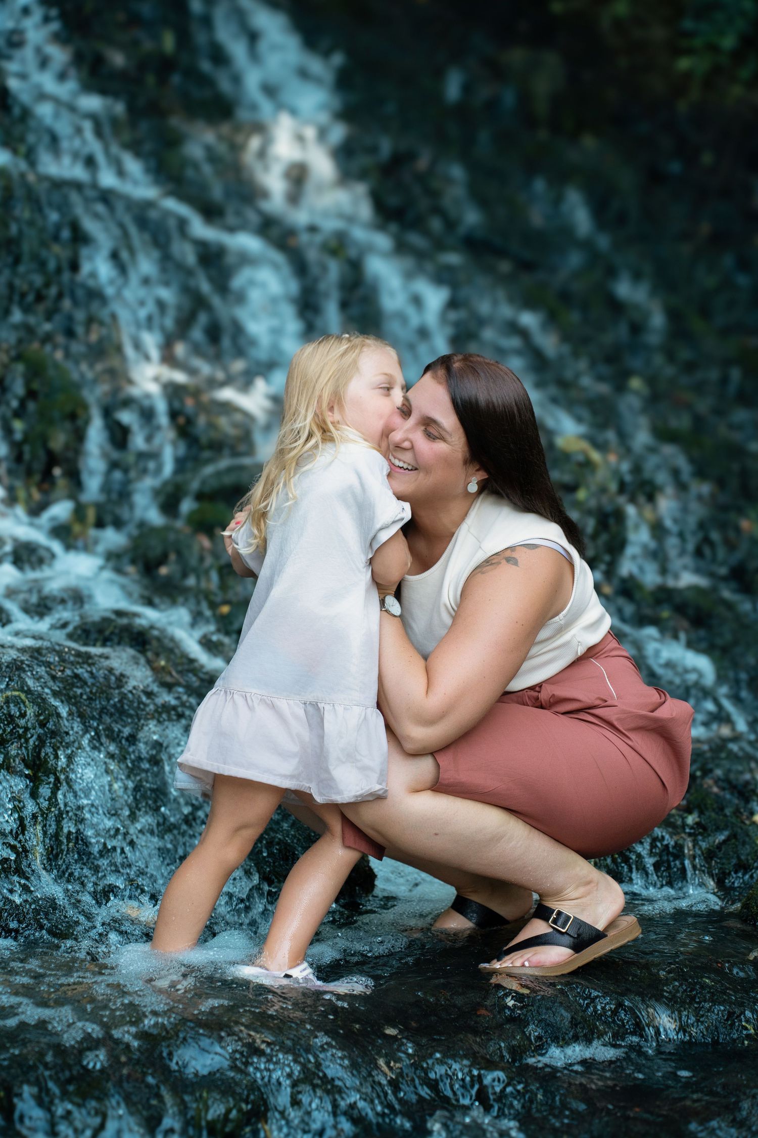 A mother crouches by a waterfall, hugging her young blonde daughter in a white dress, sharing a joyful moment.