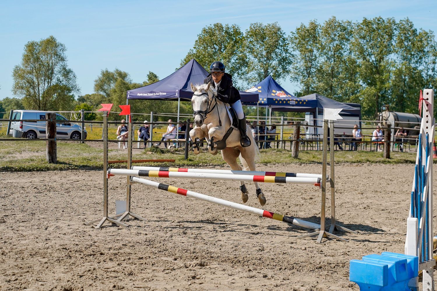 Rider on white horse jumping over colorful show jumping fence at outdoor equestrian competition.