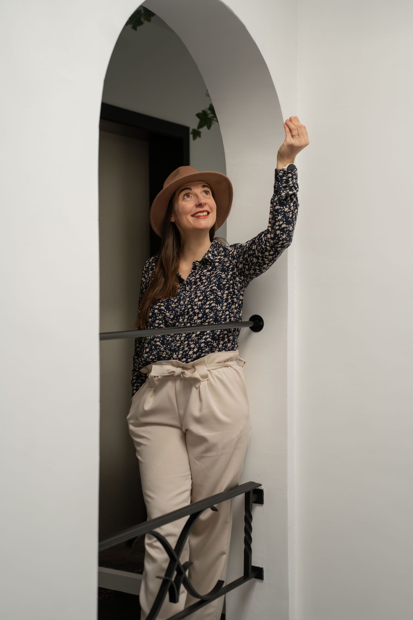 Person in hat and floral print top posing playfully under white archway.