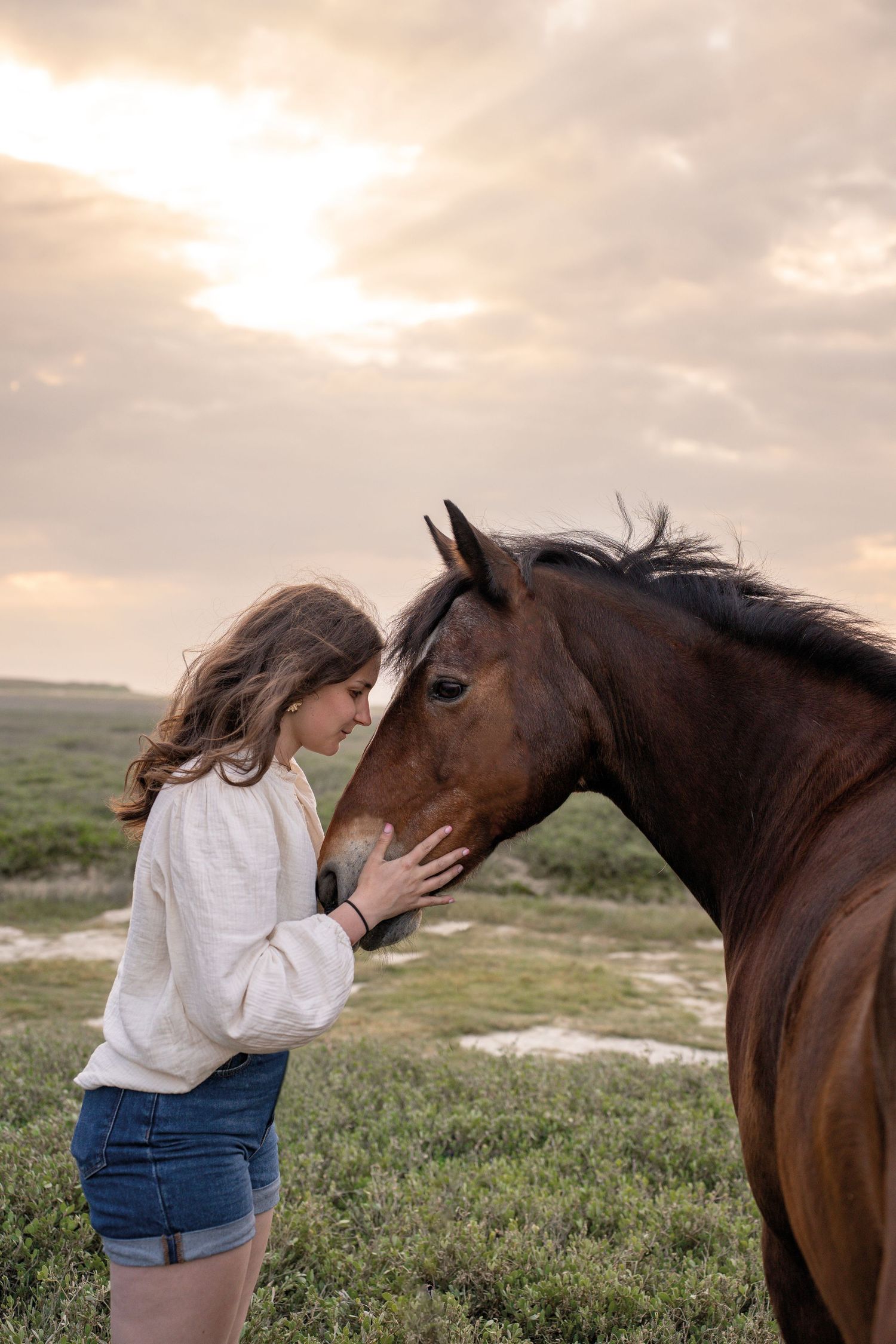 Young woman in white top tenderly touches foreheads with a bay horse at sunset in a green field.