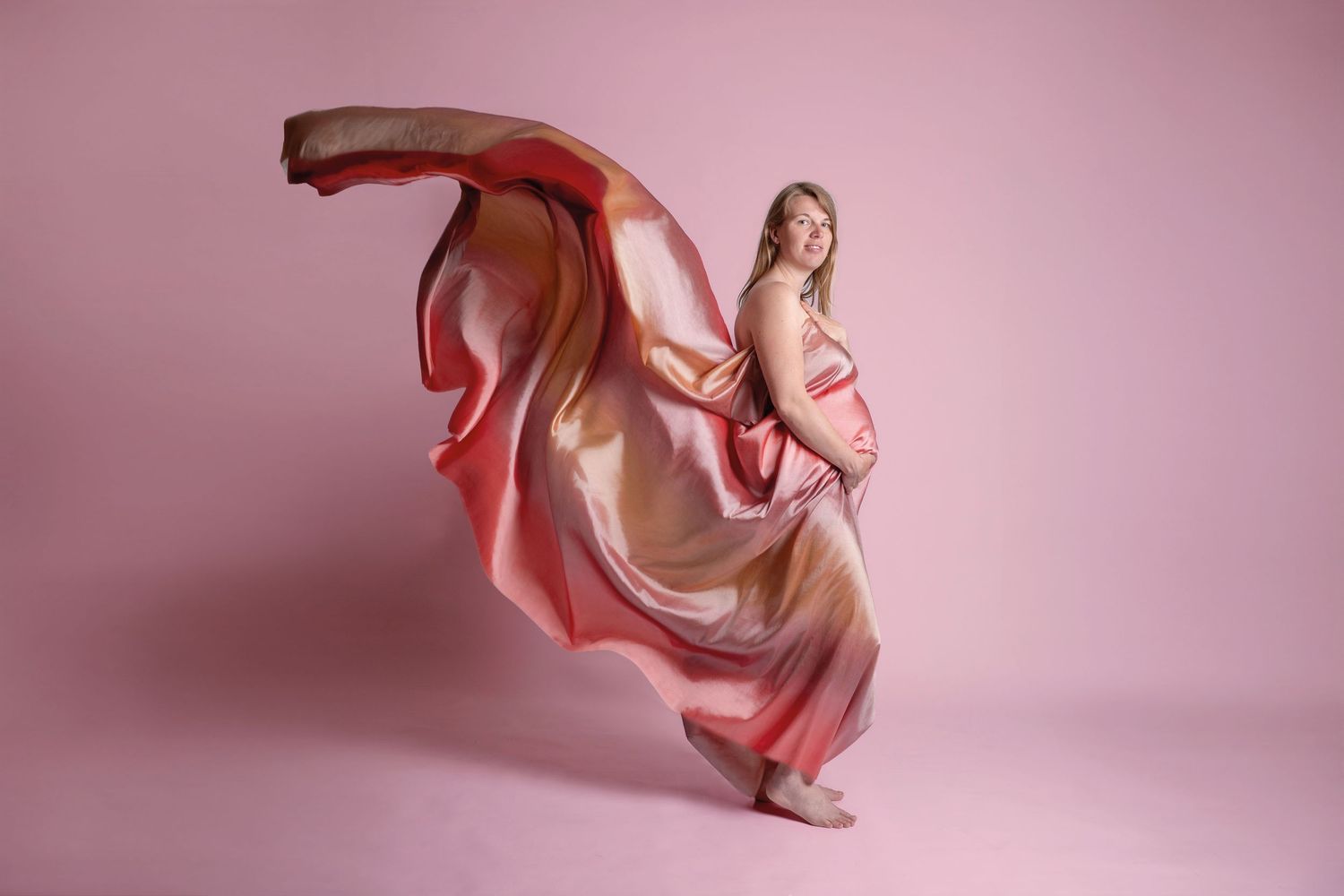 Pregnant woman in flowing pink fabric dress poses gracefully against a pink studio background.