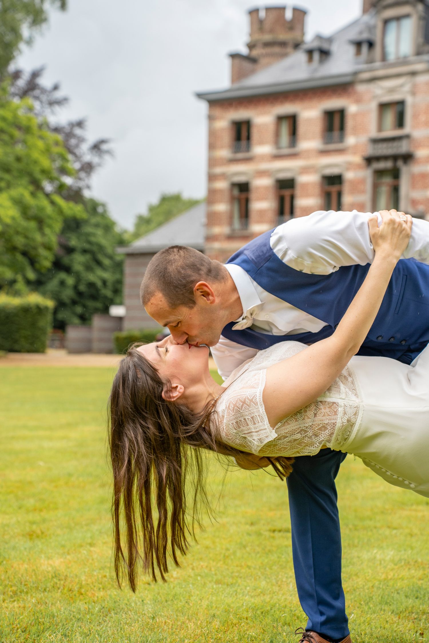 Couple performs romantic dip pose on green lawn in front of historic mansion.