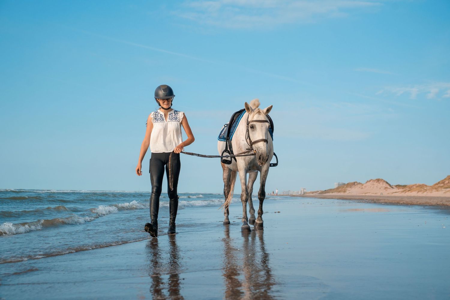 Woman in riding gear leading a white horse along a sunny beach shoreline with blue skies.