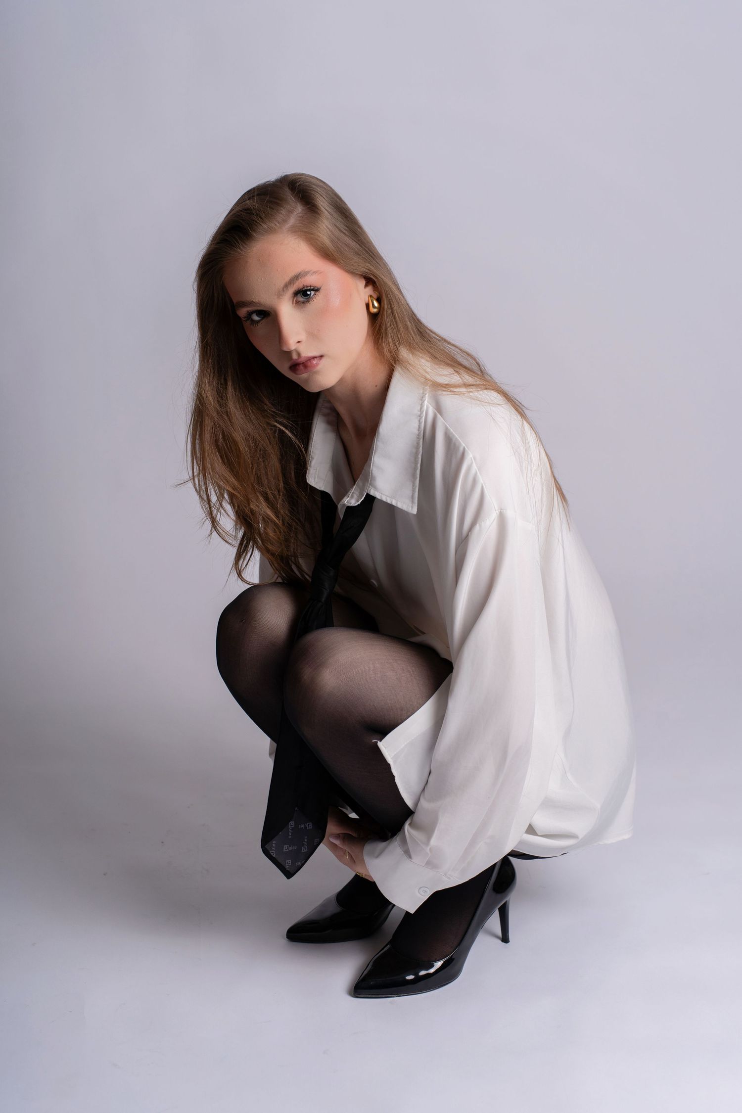 Young woman in white shirt and black tights crouching in studio, wearing patent black heels.