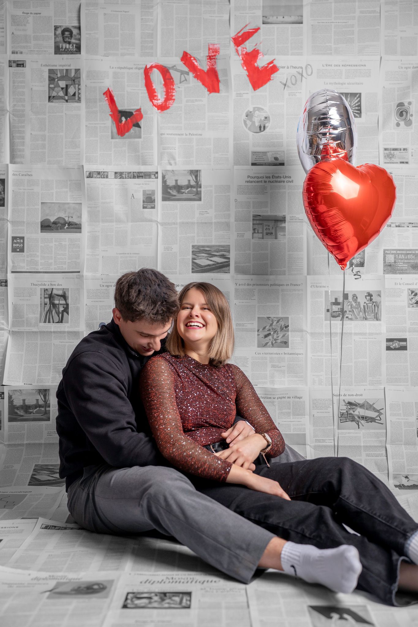 Series of romantic poses with couple lounging against newspaper wall decorated with red heart balloons.