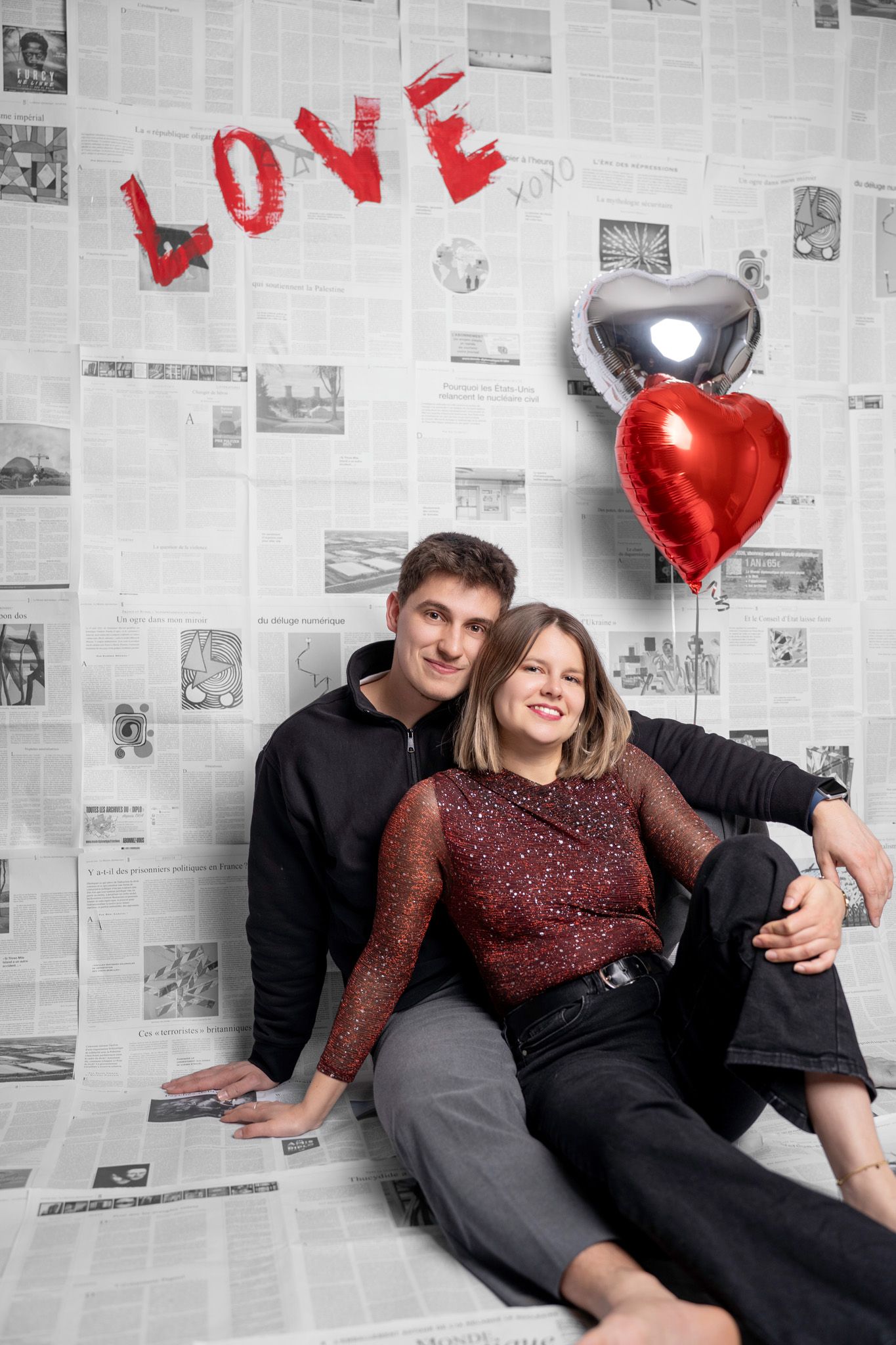 Romantic couple poses against newspaper wall with red heart balloons and 'LOVE' text in Valentine's themed photoshoot.