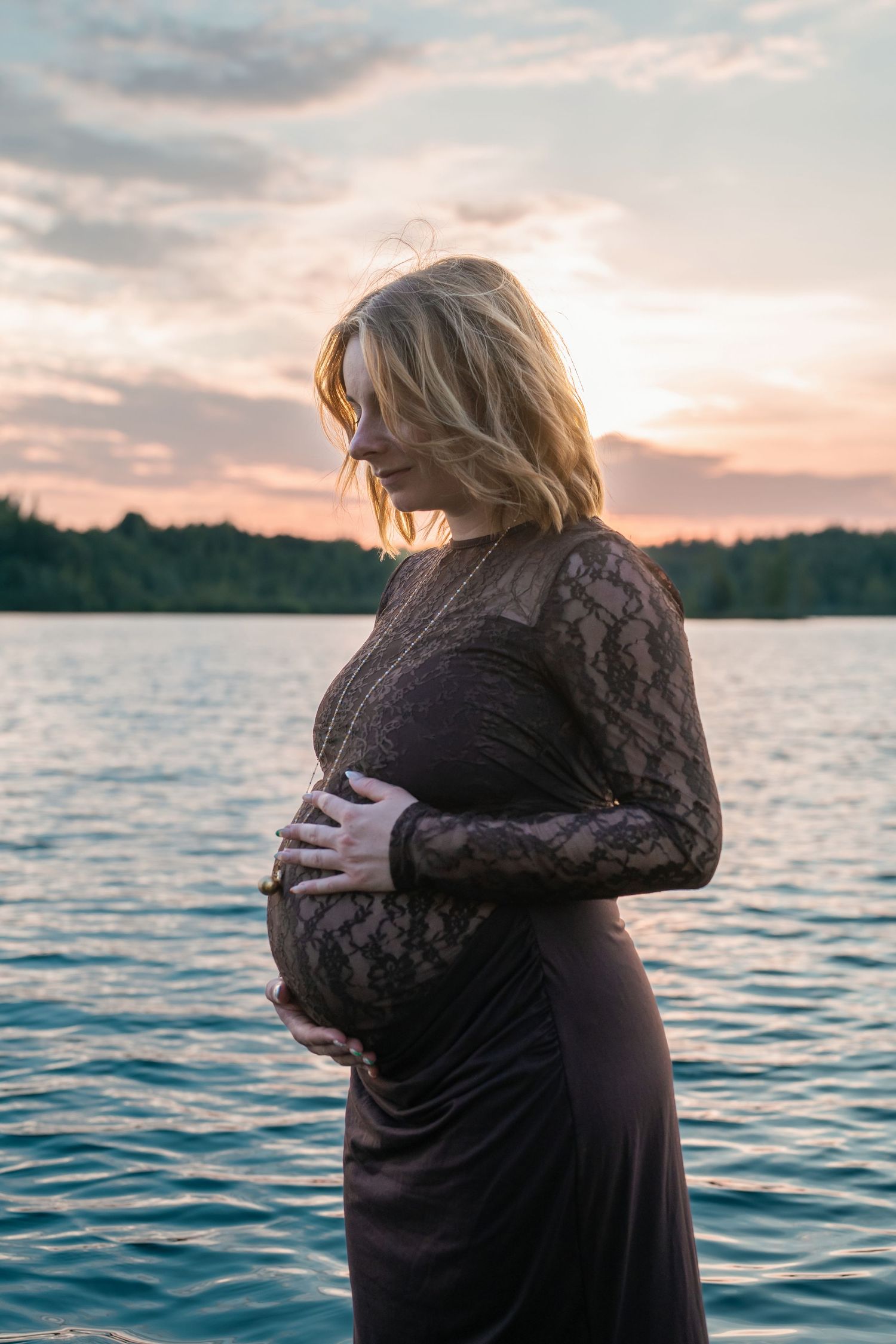 Pregnant woman in black lace dress cradling baby bump by lake at sunset.