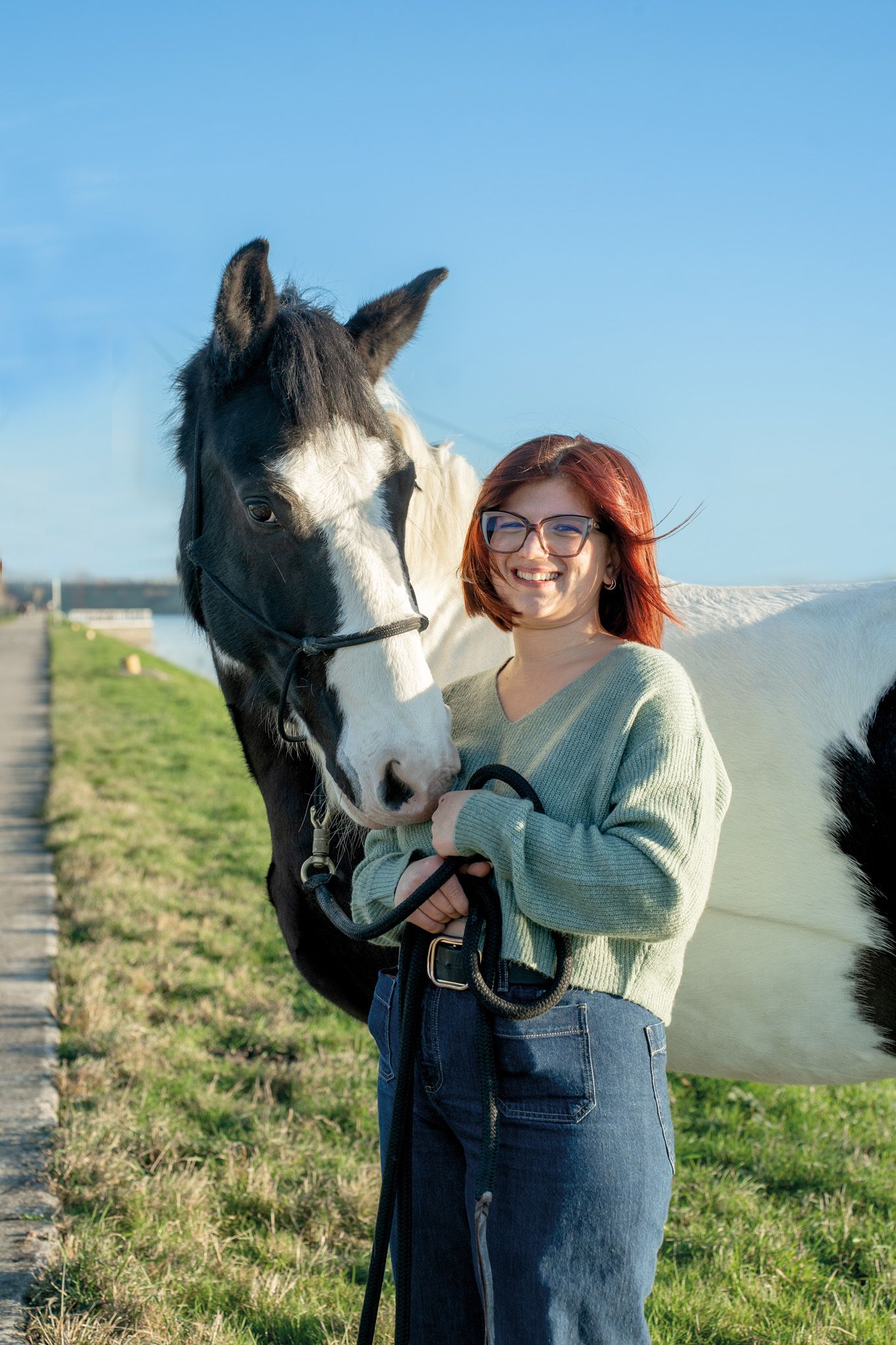 Horse and companion share tender moment on grassy hillside under blue sky.
