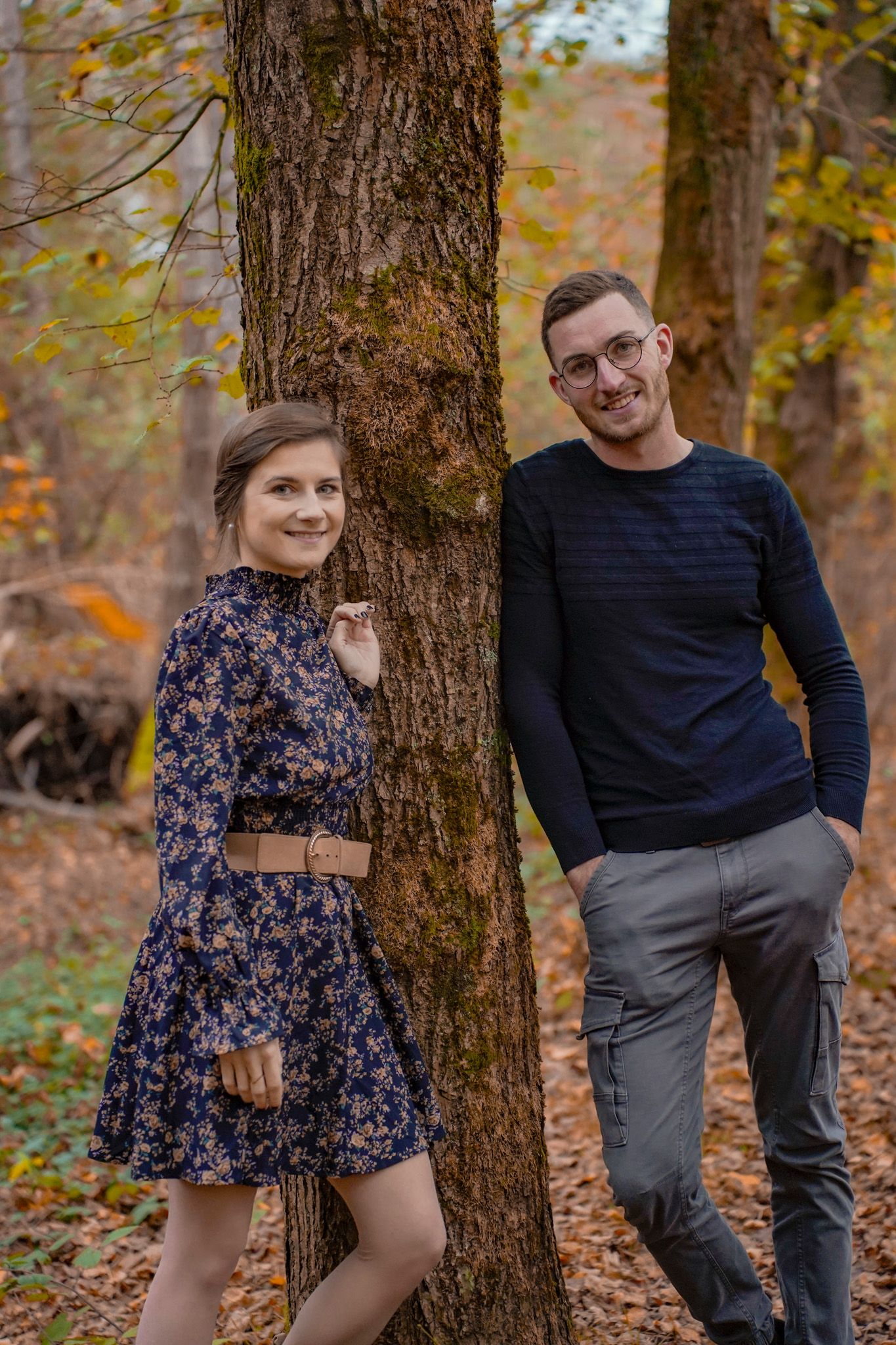 Playful autumn photoshoot with a couple posing by tree trunks in a wooded area.
