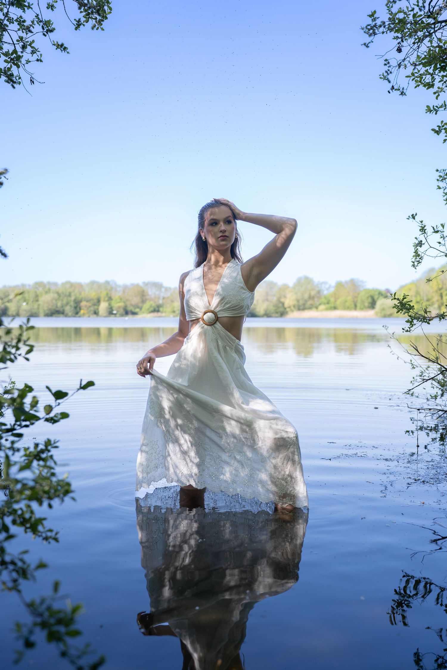 Woman in white dress standing in calm lake water, framed by trees with blue sky and green forest reflected in water.