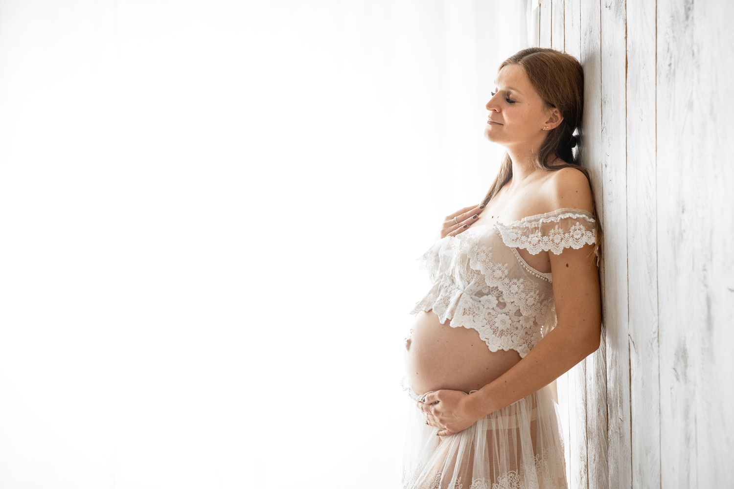 Elegant maternity portrait in white lace dress against rustic wooden wall.
