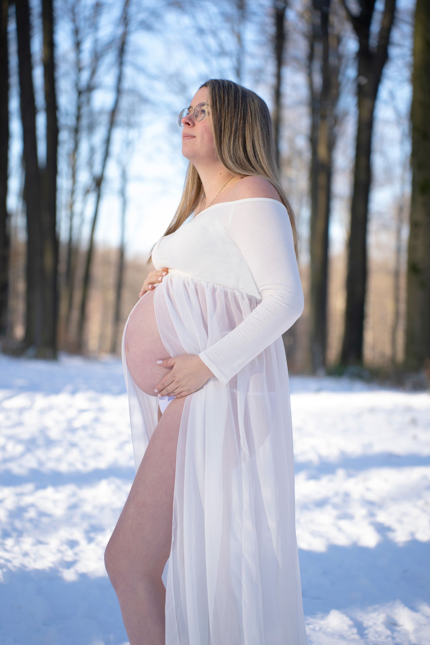Winter maternity photos featuring a white flowy dress against snowy forest backdrop.
