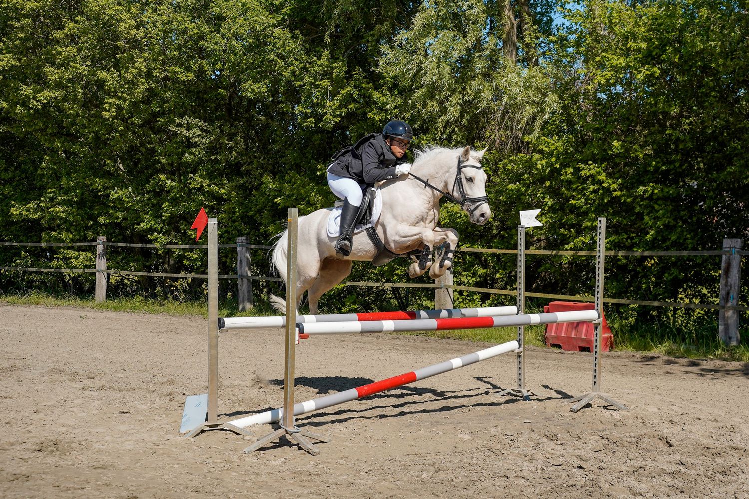 White horse and rider jumping over a red and white fence obstacle in an outdoor equestrian show jumping competition.