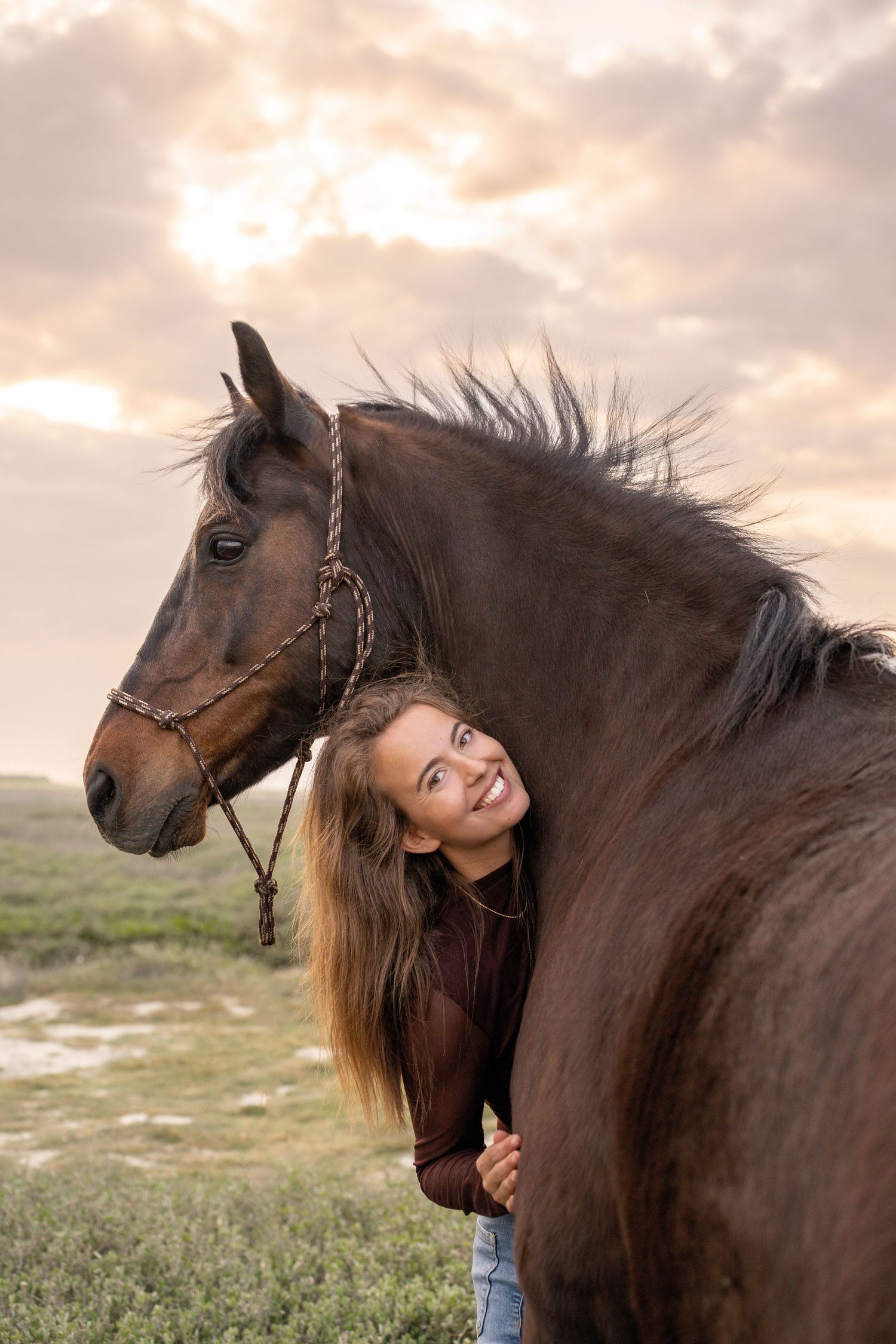 Smiling young woman with long hair peeking around a dark brown horse wearing a rope halter at sunset.
