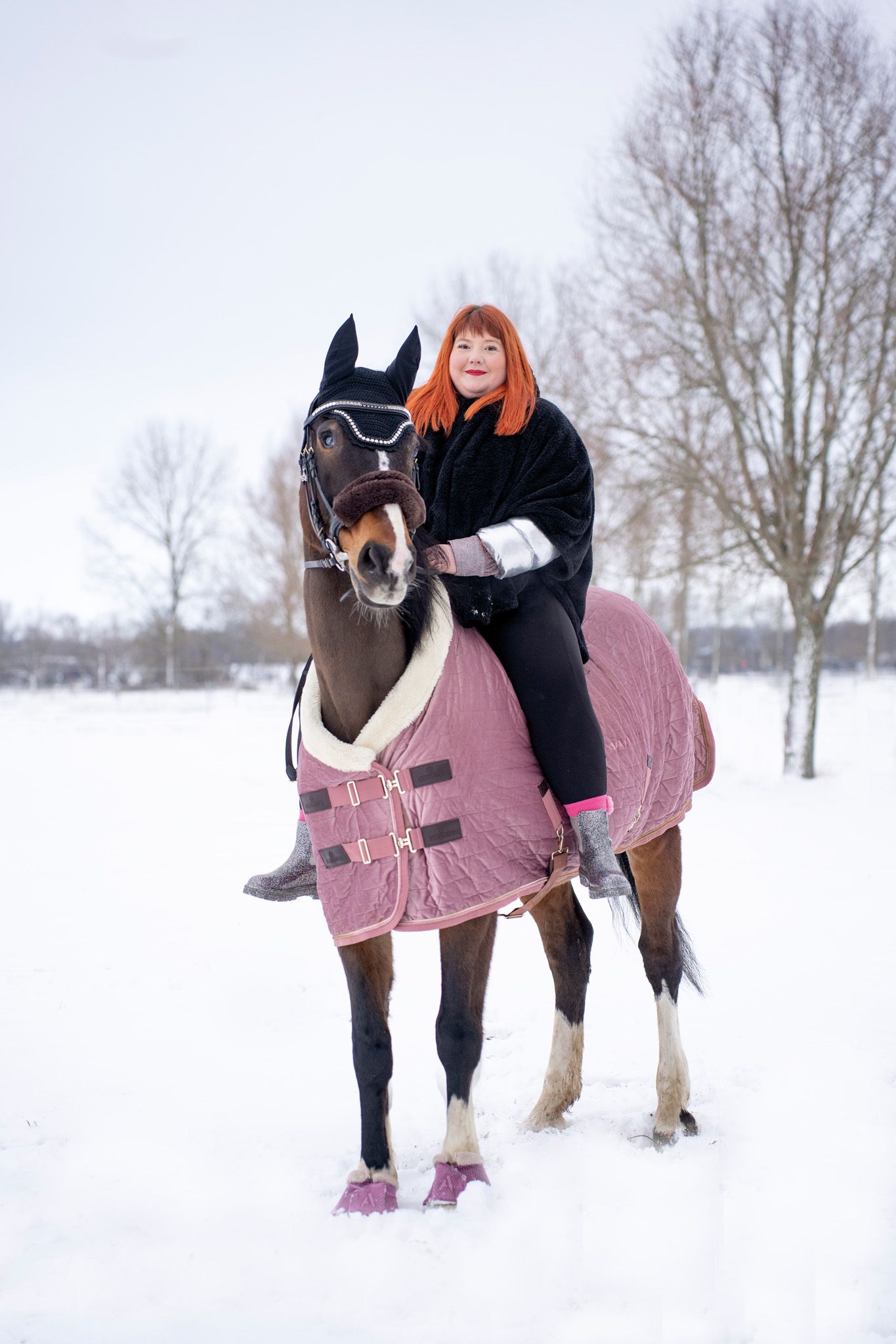 Horse wearing a pink winter blanket stands in snowy field while being cared for on a winter day.