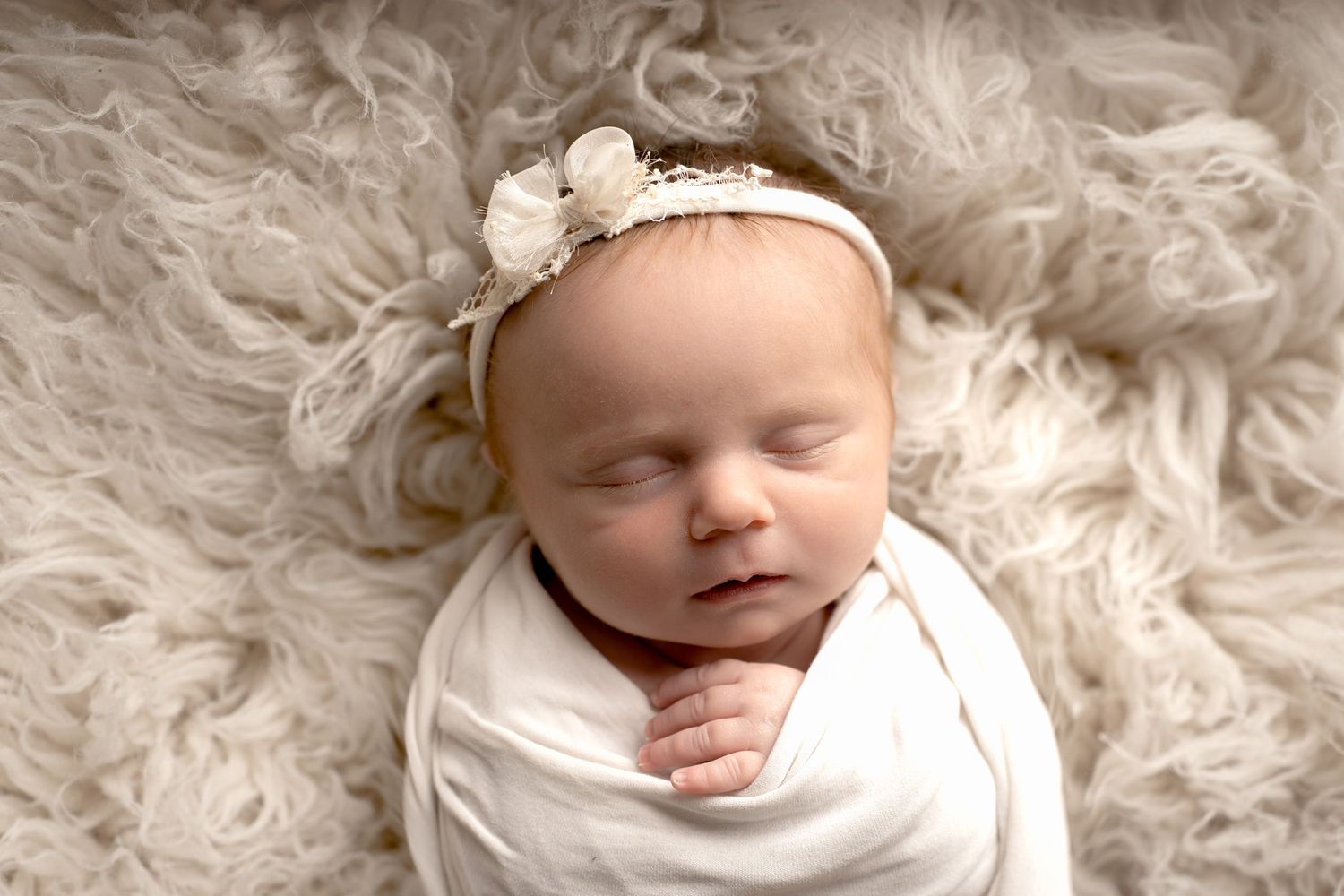 Sleeping newborn baby wrapped in white with floral headband on fluffy rug.