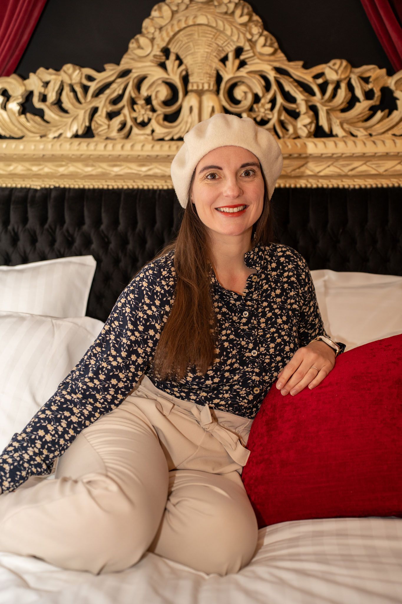 Person relaxing on a luxurious bed with red pillows and ornate gold headboard.
