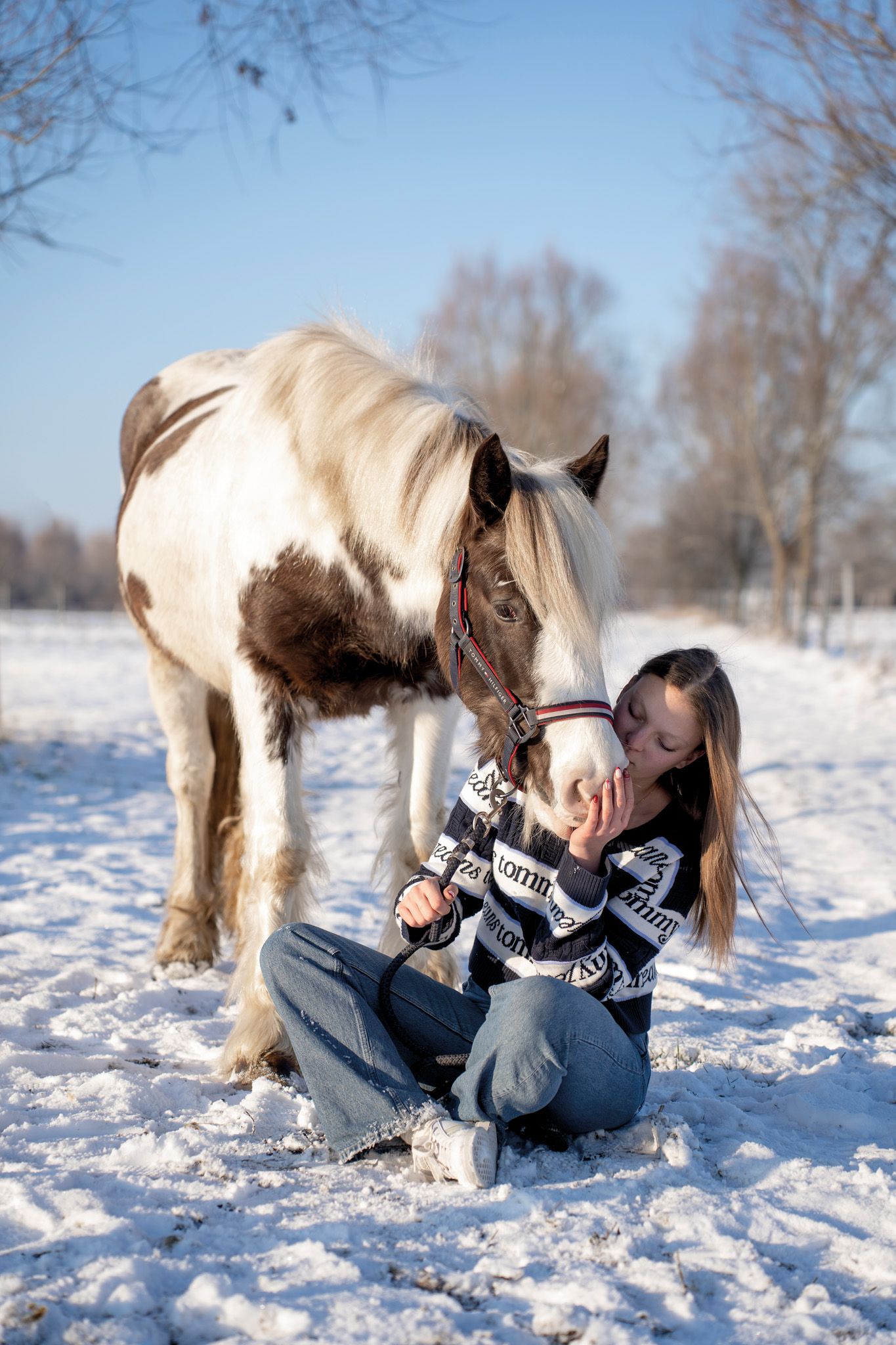 A white horse nuzzles someone lying in fresh snow during a winter photoshoot.