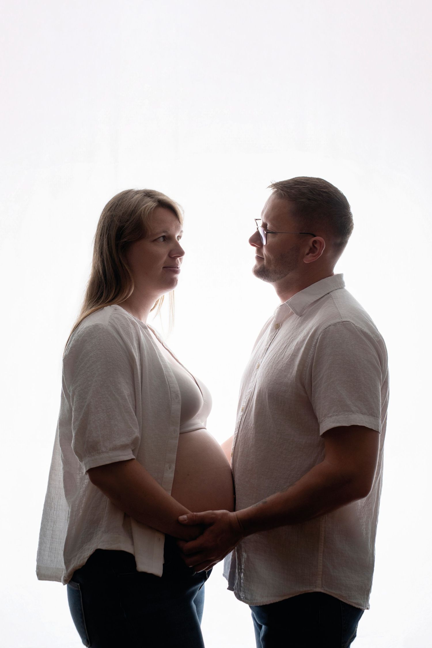 Pregnant couple holding hands, facing each other in a tender moment, backlit against a white background.