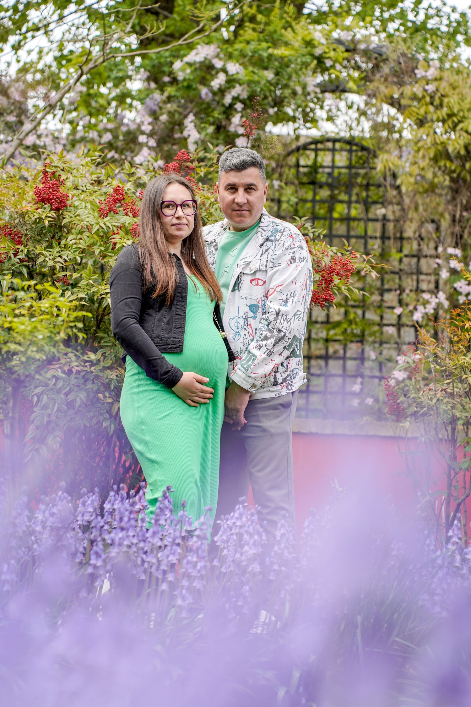 Expectant couple poses in a blooming garden with spring flowers and a decorative trellis arch.