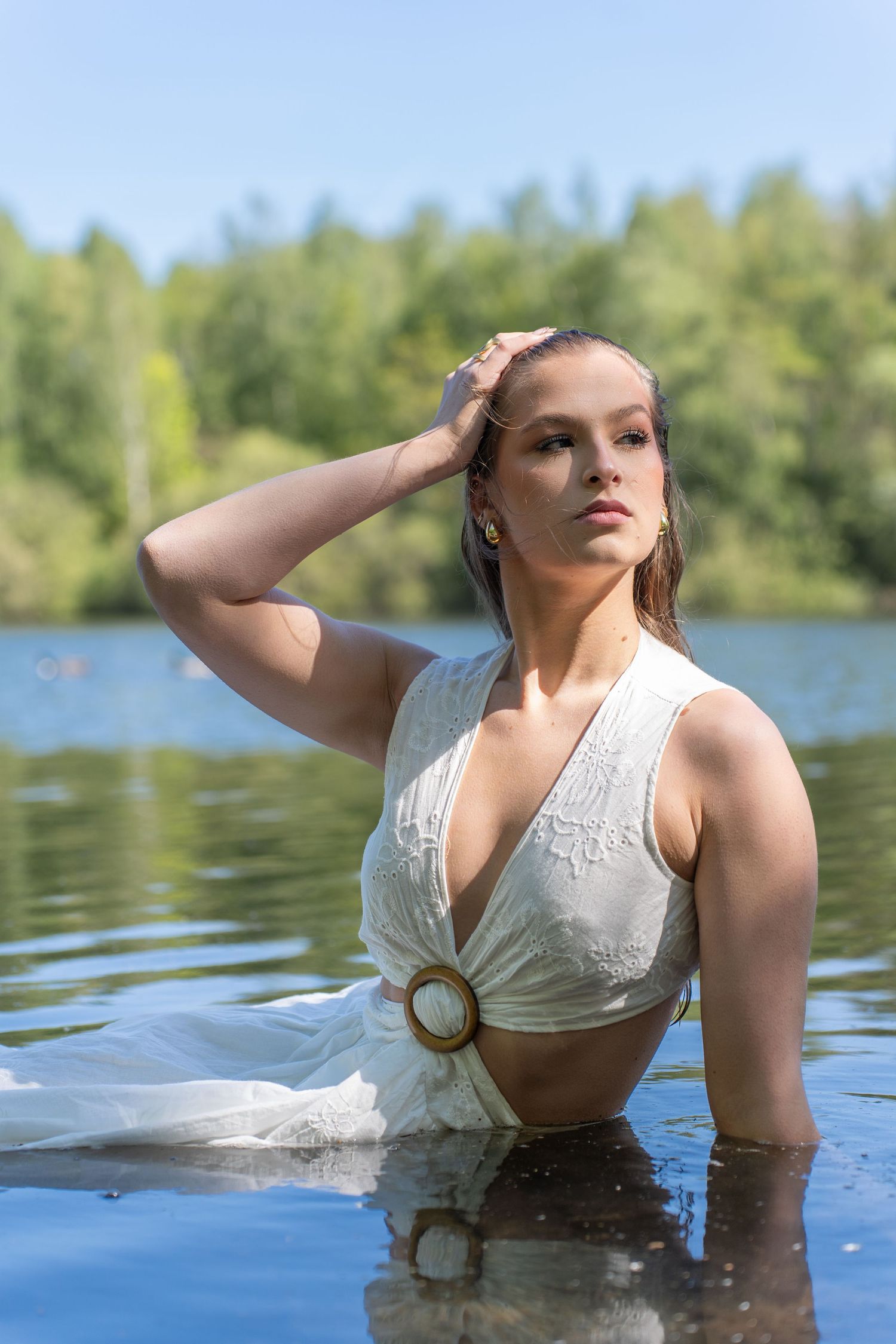 Young woman in white dress standing in a lake, hand on head, with lush green trees in the background.