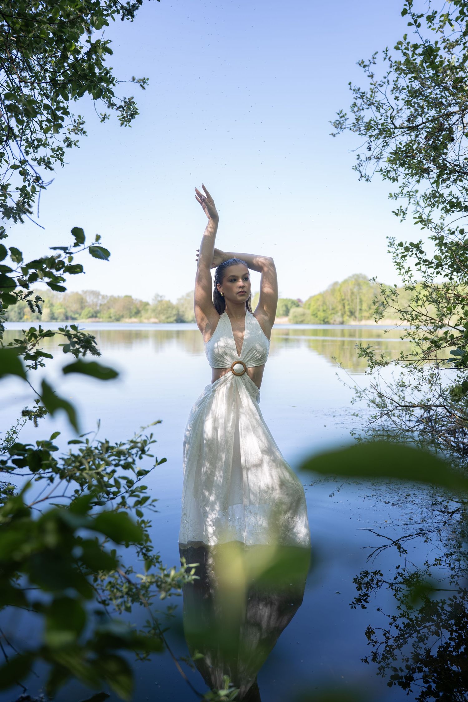 Woman in white halter dress standing in a lake, arms raised gracefully, framed by lush green trees under a clear blue sky.