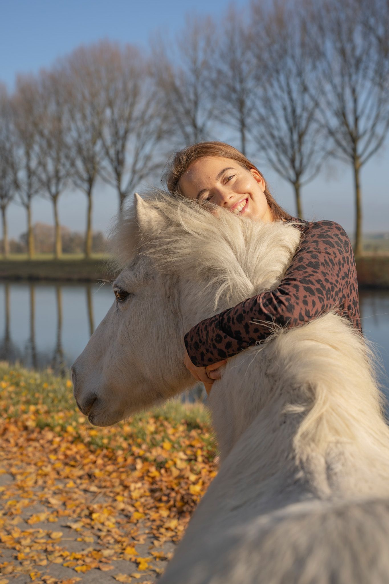 White horse with flowing mane being embraced in golden autumn sunlight by water.