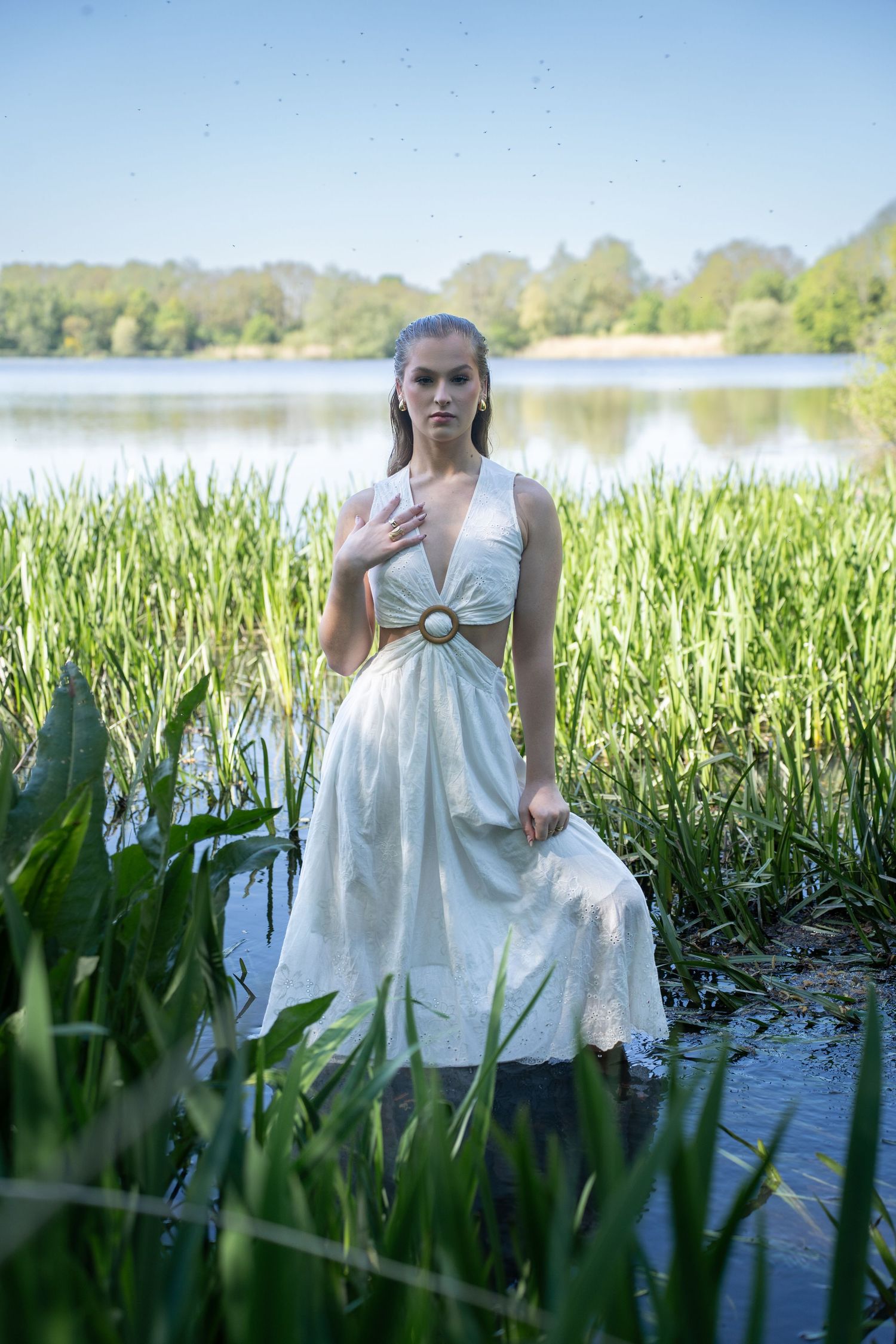 Young woman in white dress standing in lake surrounded by green reeds on a sunny day.