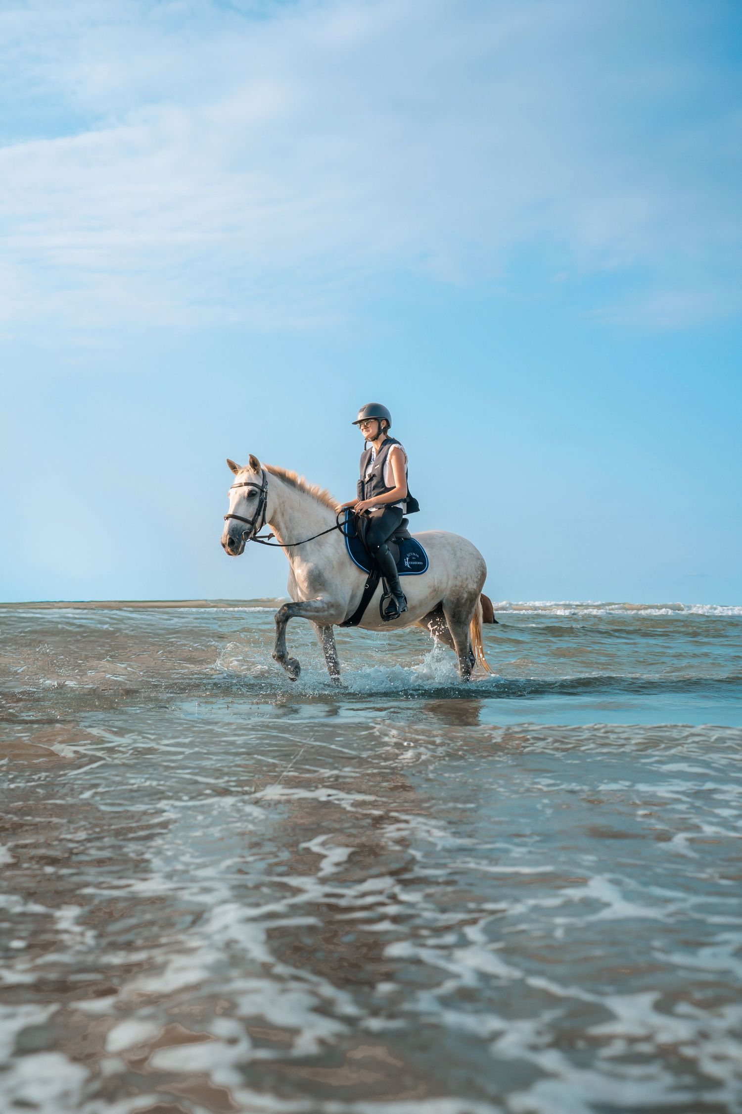 A rider on a white horse walks through shallow ocean waves on a beach under a clear blue sky.