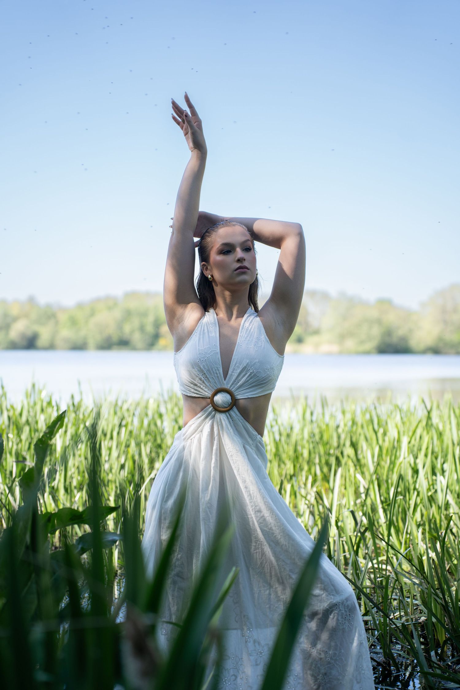 Woman in white halter dress poses gracefully among tall reeds by a serene lake on a sunny day.