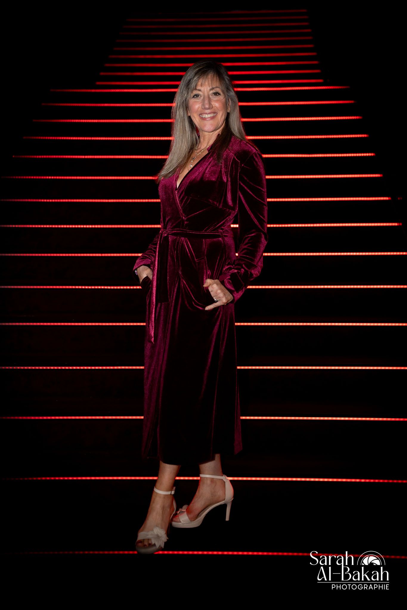 Elegant portrait on red-lit stairs wearing a burgundy velvet dress.