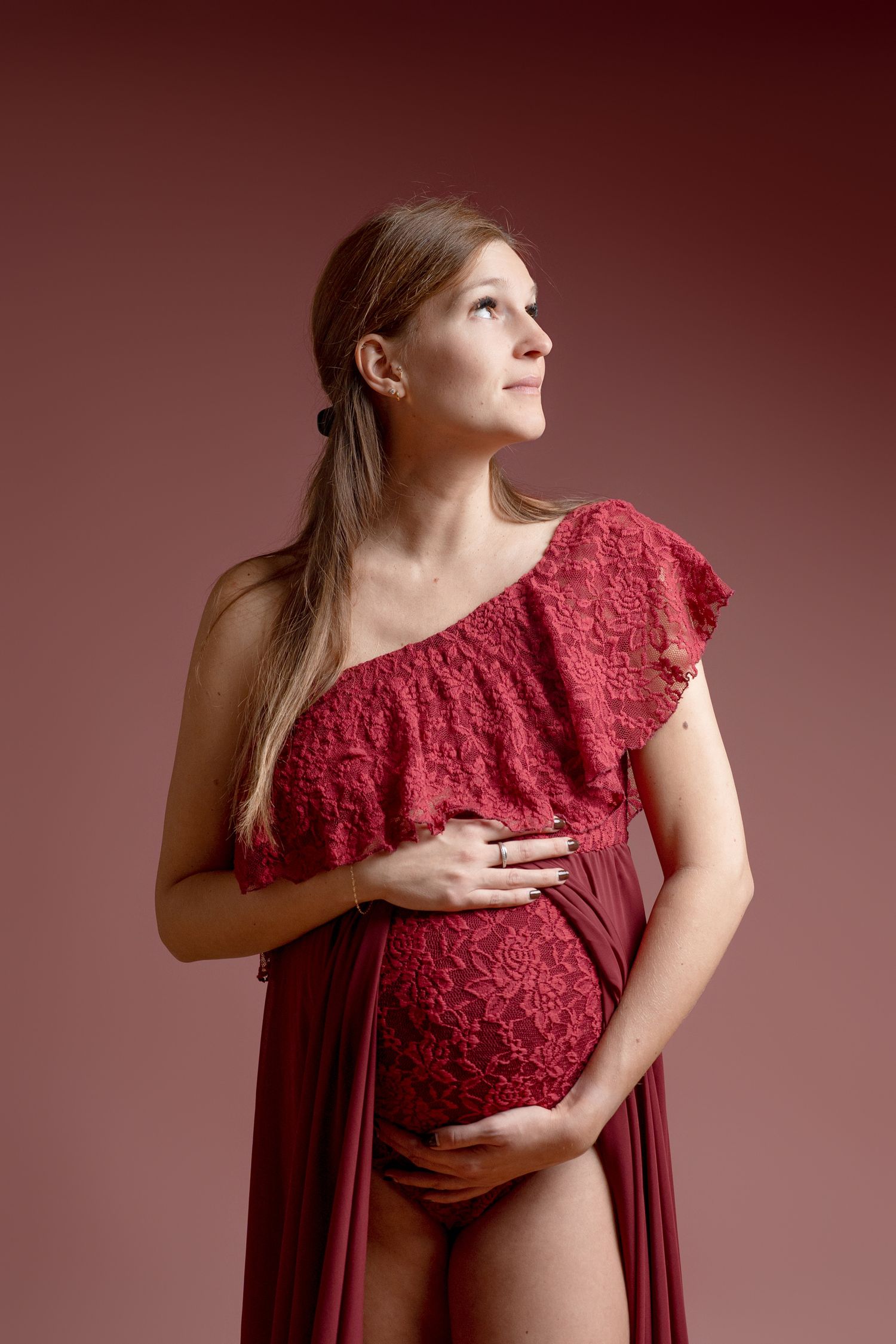 Elegant maternity portraits in flowing red lace dress against burgundy backdrop.