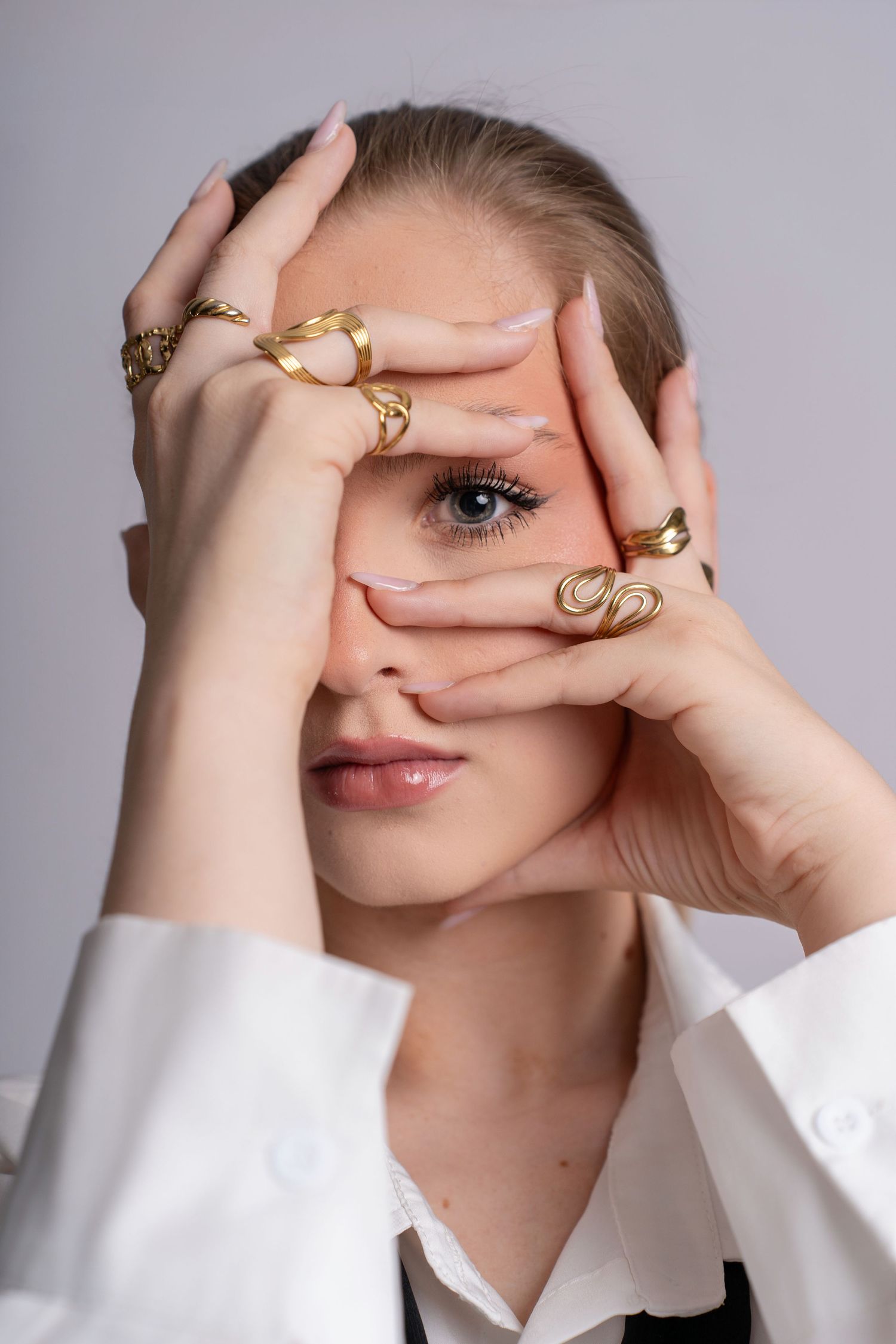 Woman wearing multiple gold rings on both hands, framing her blue eye while wearing a white shirt.