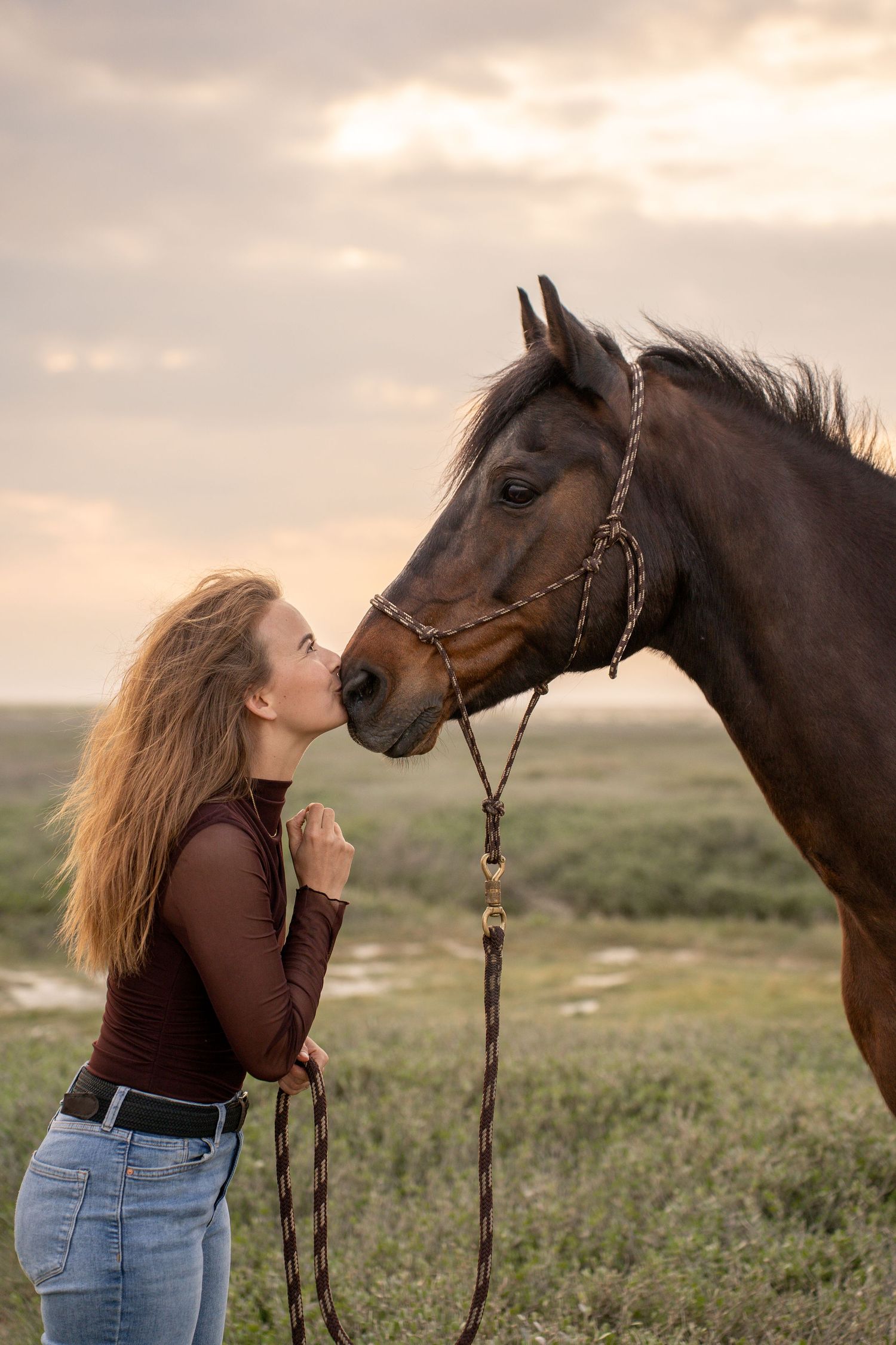Young woman with long hair kissing a brown horse wearing a rope halter in an open field at sunset.