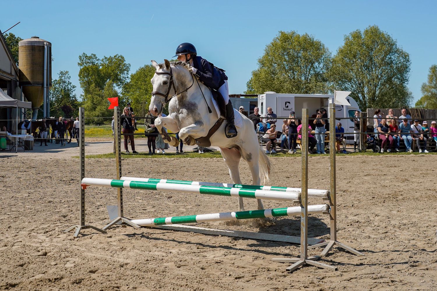 A rider on a white horse jumping over a green and white fence obstacle at an outdoor equestrian show.