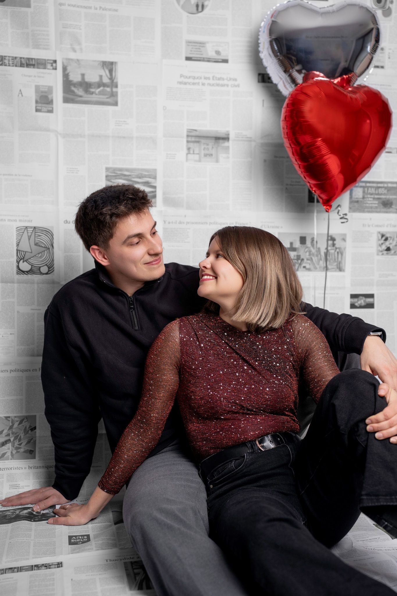 Sweet couple relaxes on floor with heart balloons against newspaper wall in romantic photoshoot.