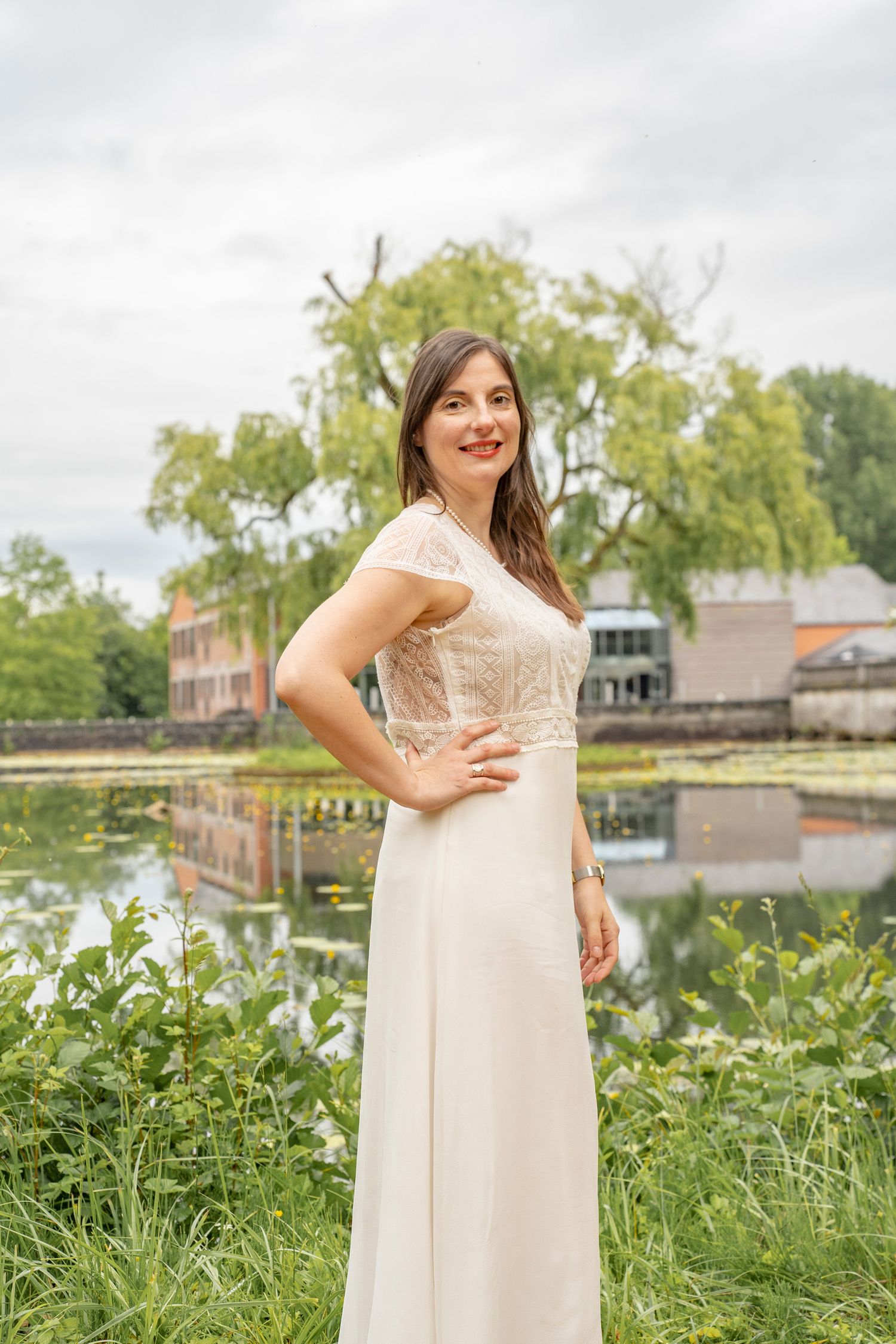 A person in a cream-colored dress poses by a lake with historic buildings in the background.