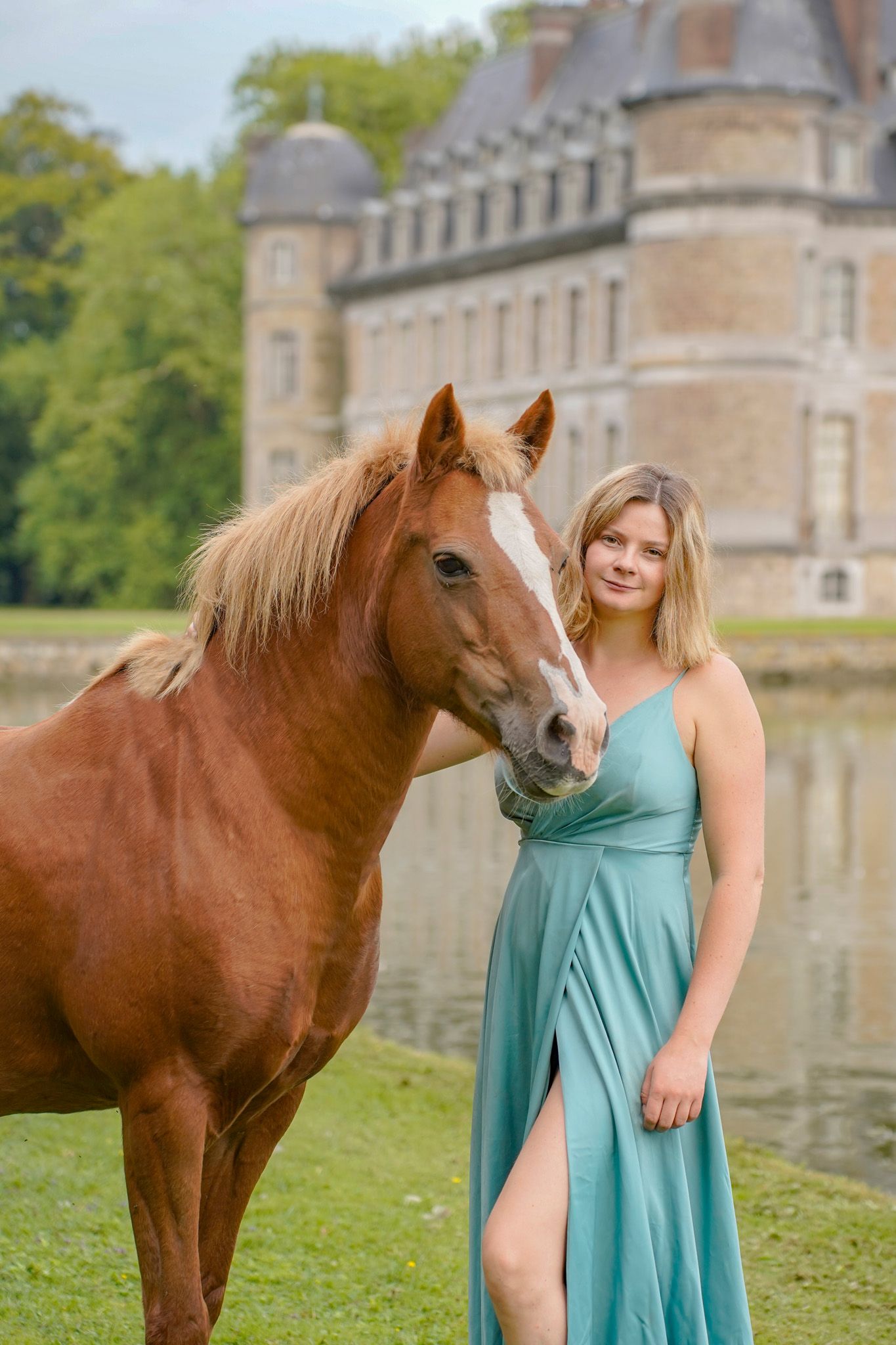 Chestnut horse posing near a grand castle with reflective pond in background.